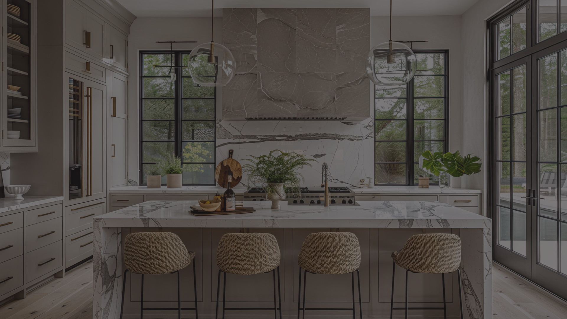 Modern kitchen with white cabinetry, marble countertops and backsplash, black window frames, and a large marble island with four woven bar stools. There are hanging pendant lights, potted plants, and sliding glass doors leading outside.