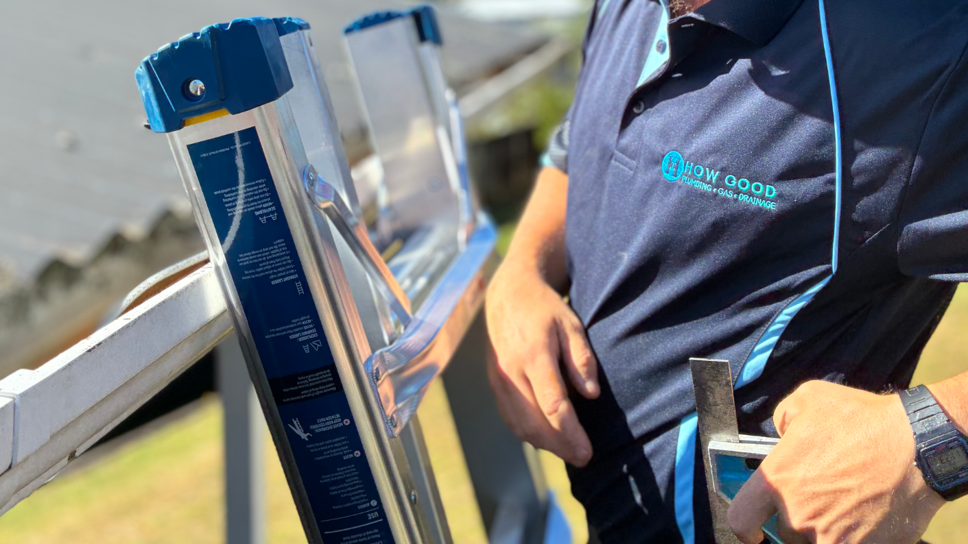 How Good plumber in a navy branded work shirt standing beside a ladder on a job