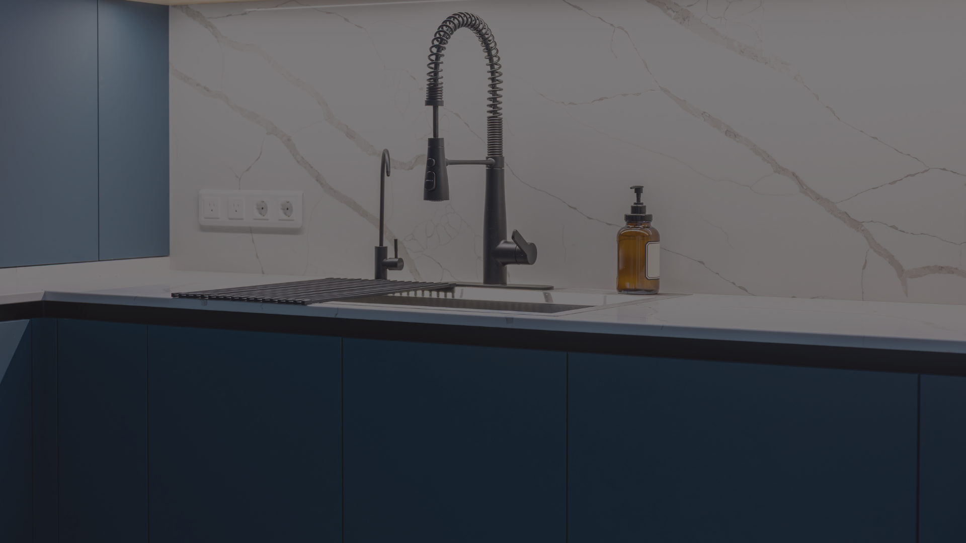 Modern kitchen sink with black faucet, brown soap dispenser, and blue cabinets, against a white marble backsplash.