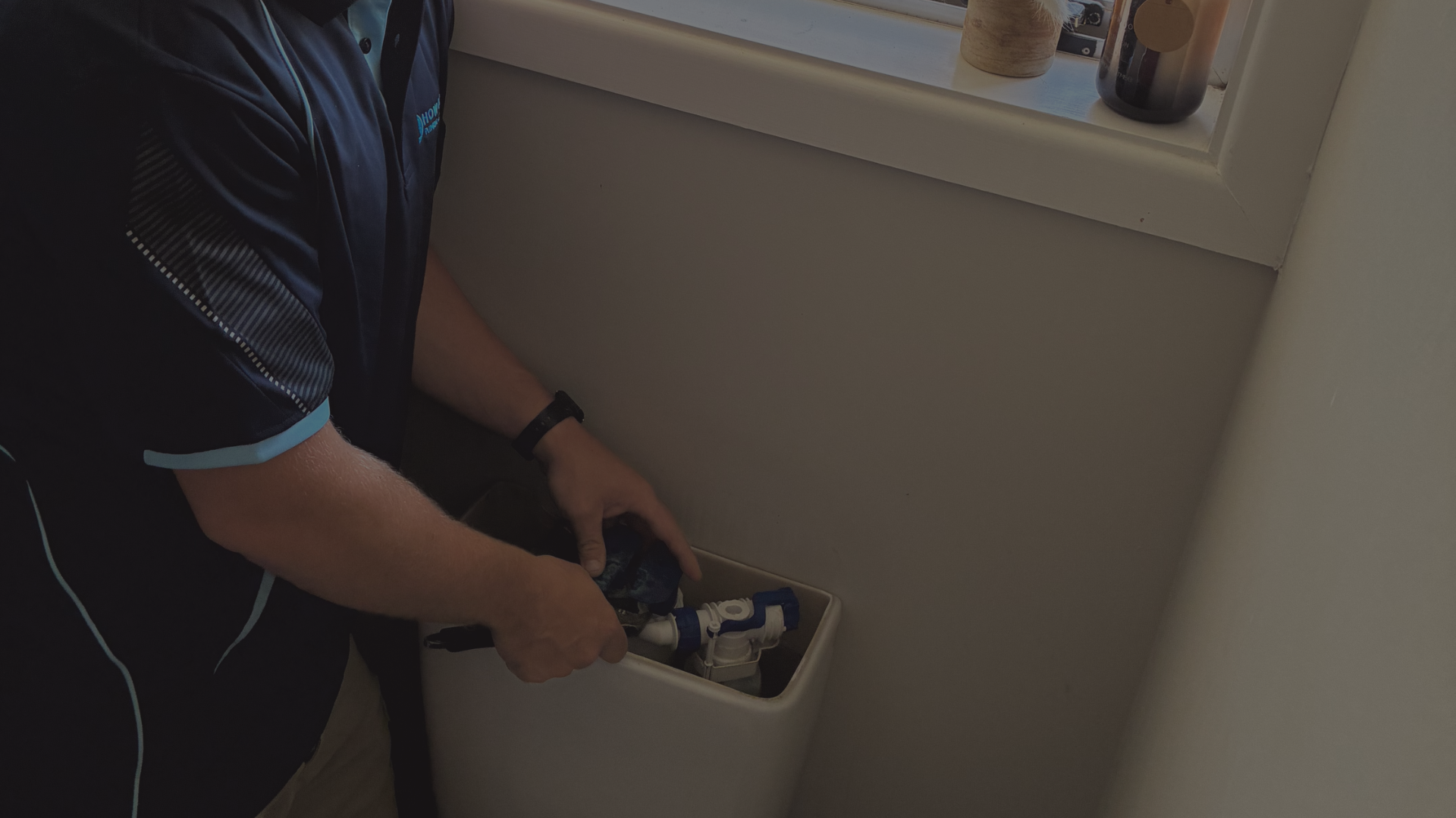 A person working on a toilet tank, possibly repairing or inspecting plumbing inside the tank near a window with decorative items.