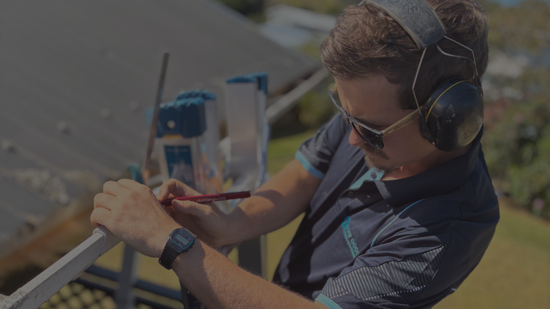 A man wearing safety headphones and sunglasses, writing on a piece of paper attached to a railing outdoors on a sunny day.