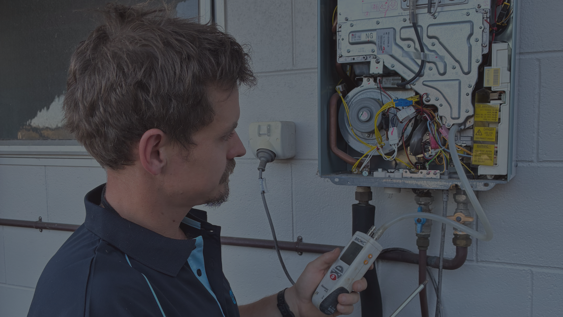 A technician repairs a gas furnace, using a digital electronic testing device, mounted on the wall in a utility room.