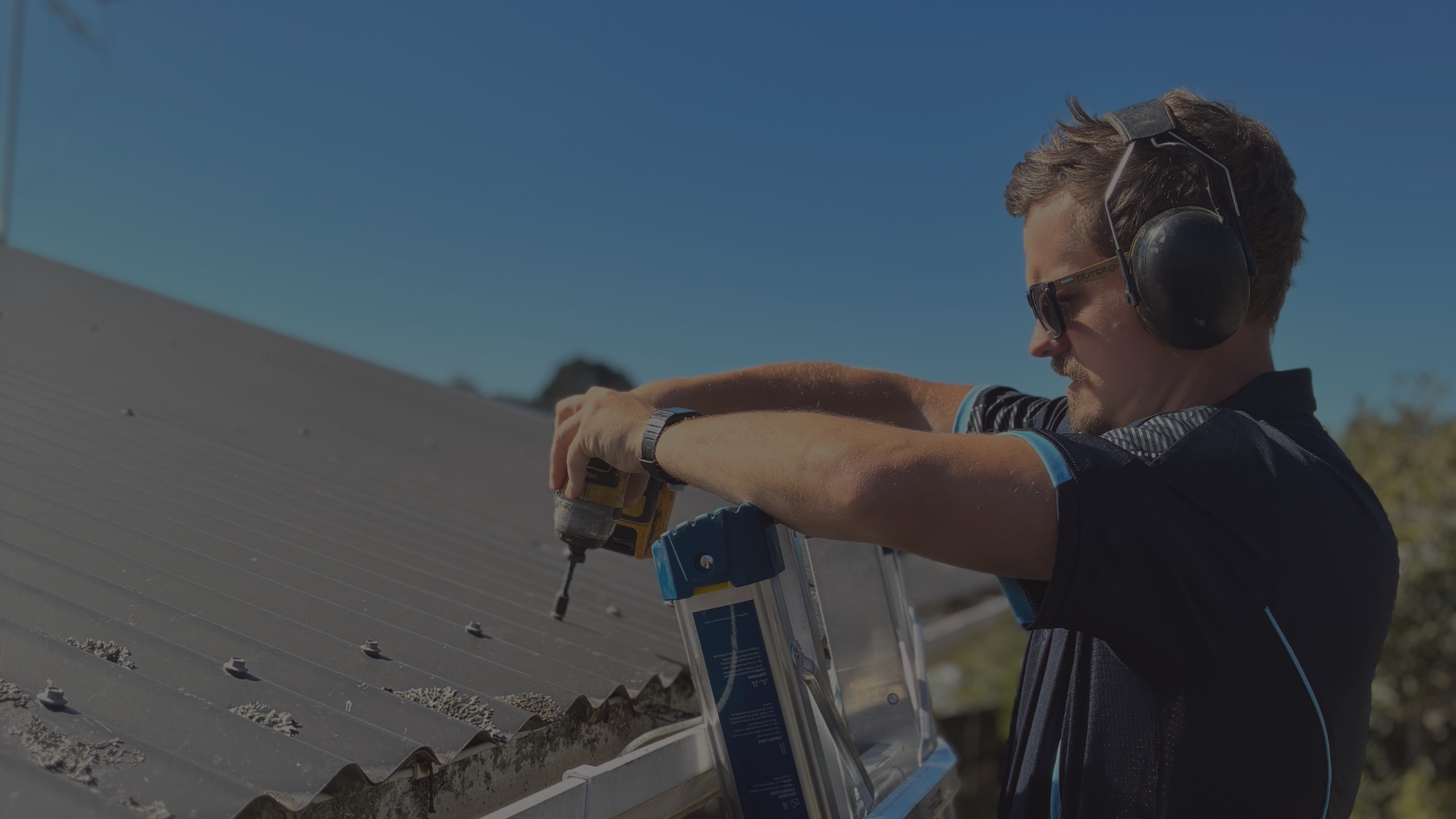 A man wearing sunglasses and work headphones is installing solar panels on a roof using a cordless drill.