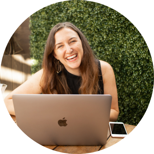 A woman with long brown hair and hoop earrings, smiling while sitting at a wooden table with a silver MacBook and smartphone, outdoors with green bushes in the background.