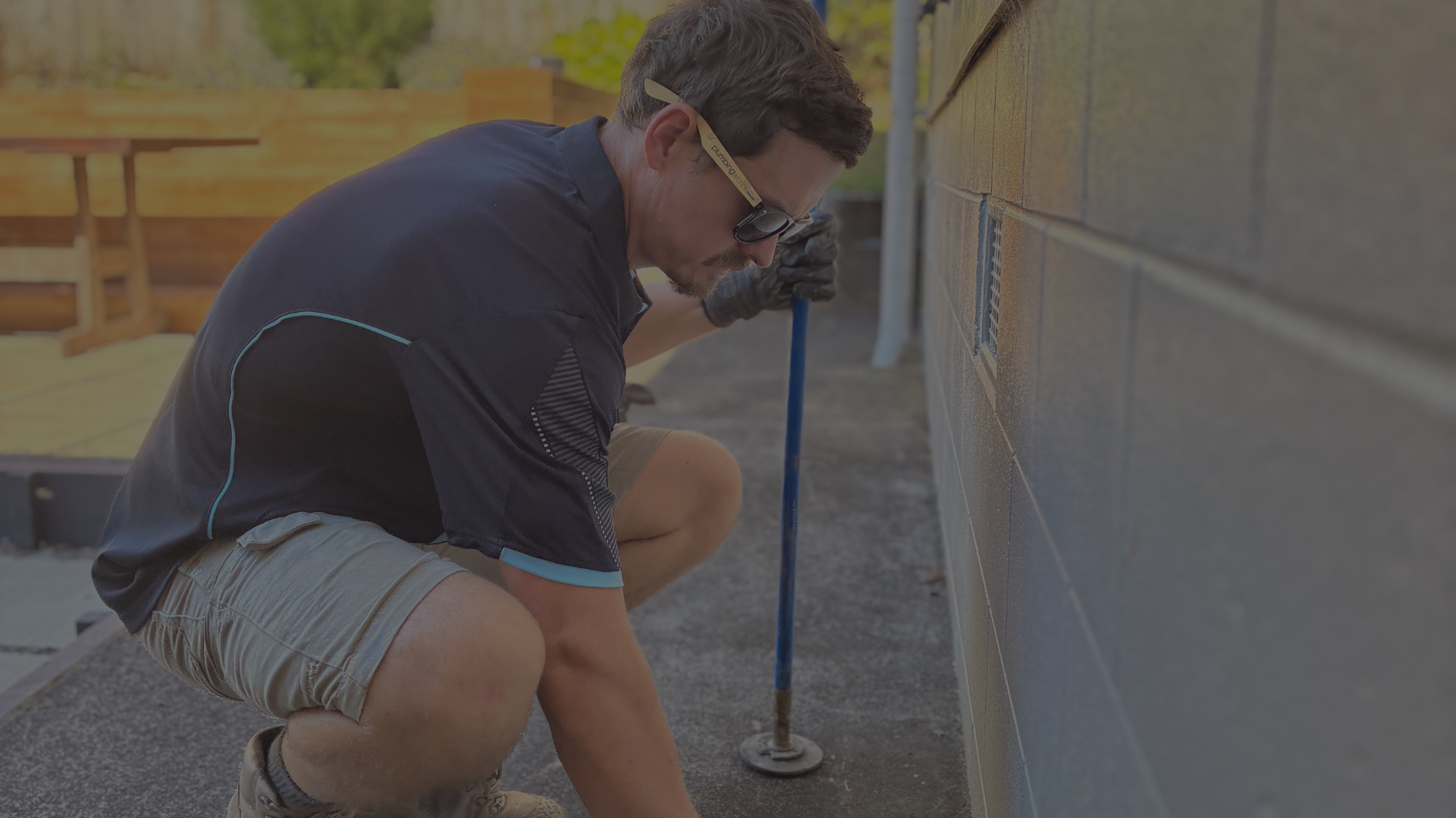 Man kneeling outdoors, working on a wall with a power tool, wearing safety glasses, black gloves, a dark shirt, and khaki shorts.