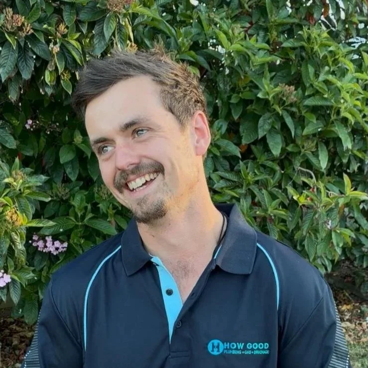 A smiling man with dark hair and a beard, wearing a navy blue polo shirt with light blue accents and a logo, standing outdoors in front of green leafy bushes.