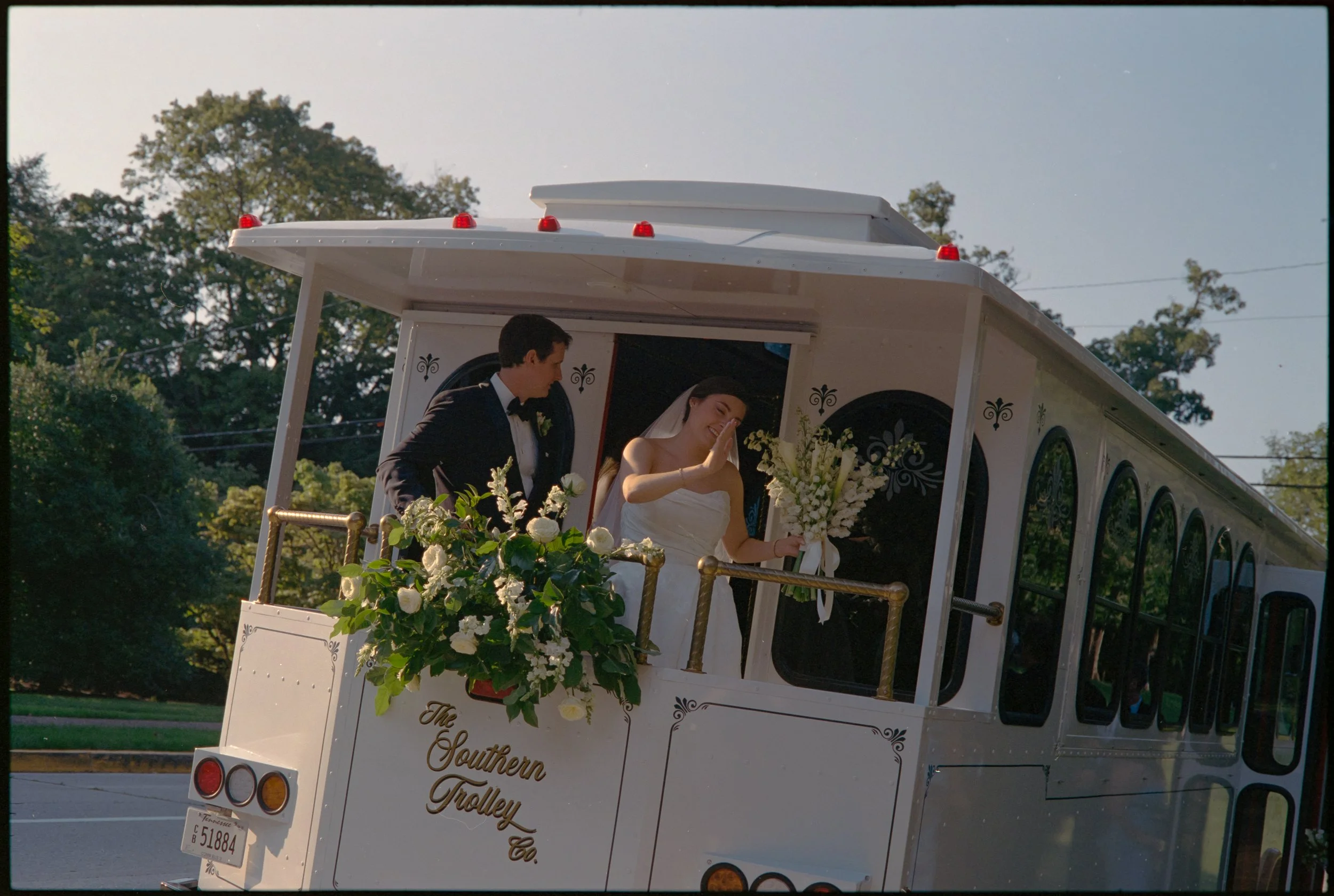 Documentary wedding photo shot on 35mm film of a couple waving to their guests from the back of a trolley