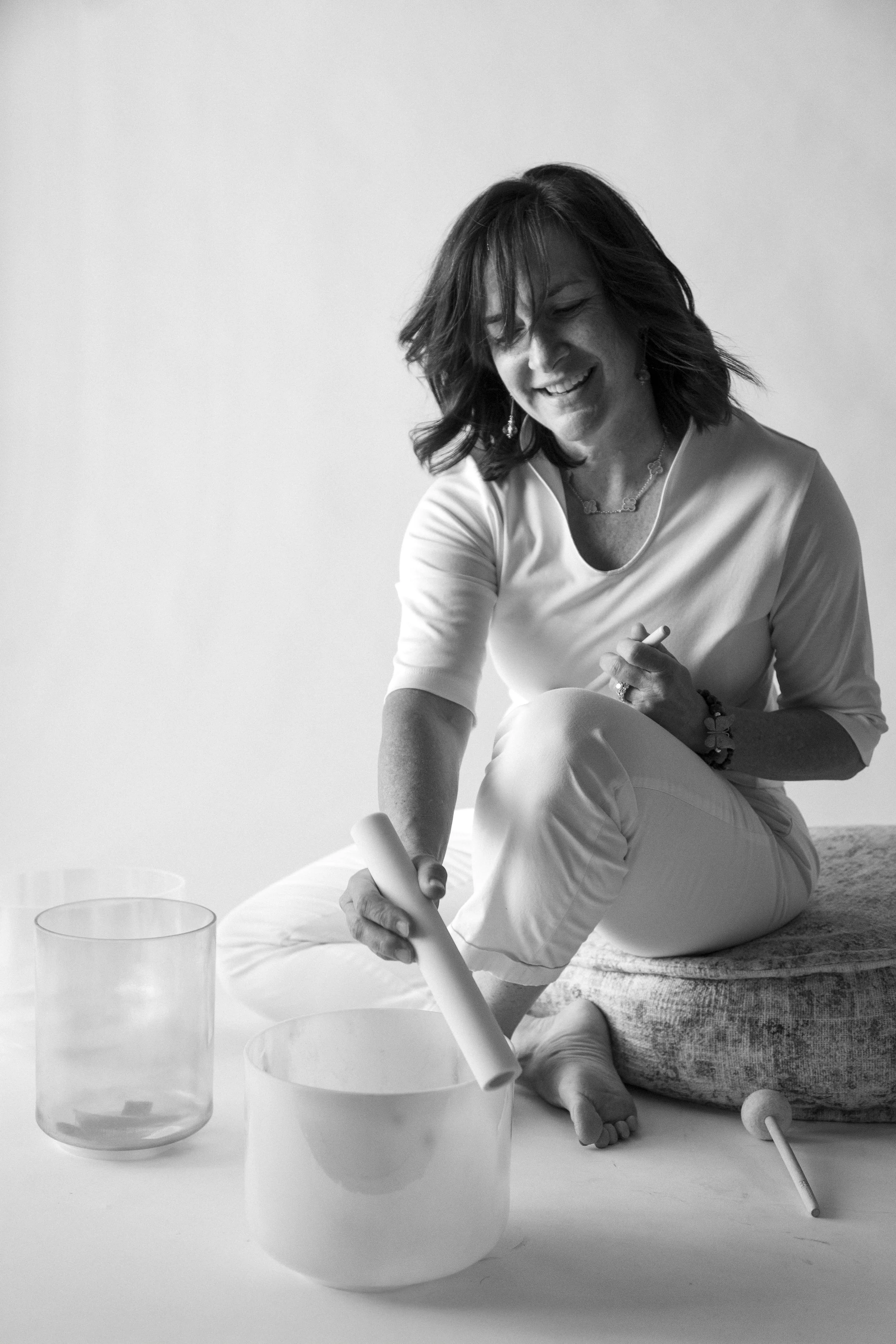 A woman sitting on a cushion, smiling at the camera, playing a singing bowl