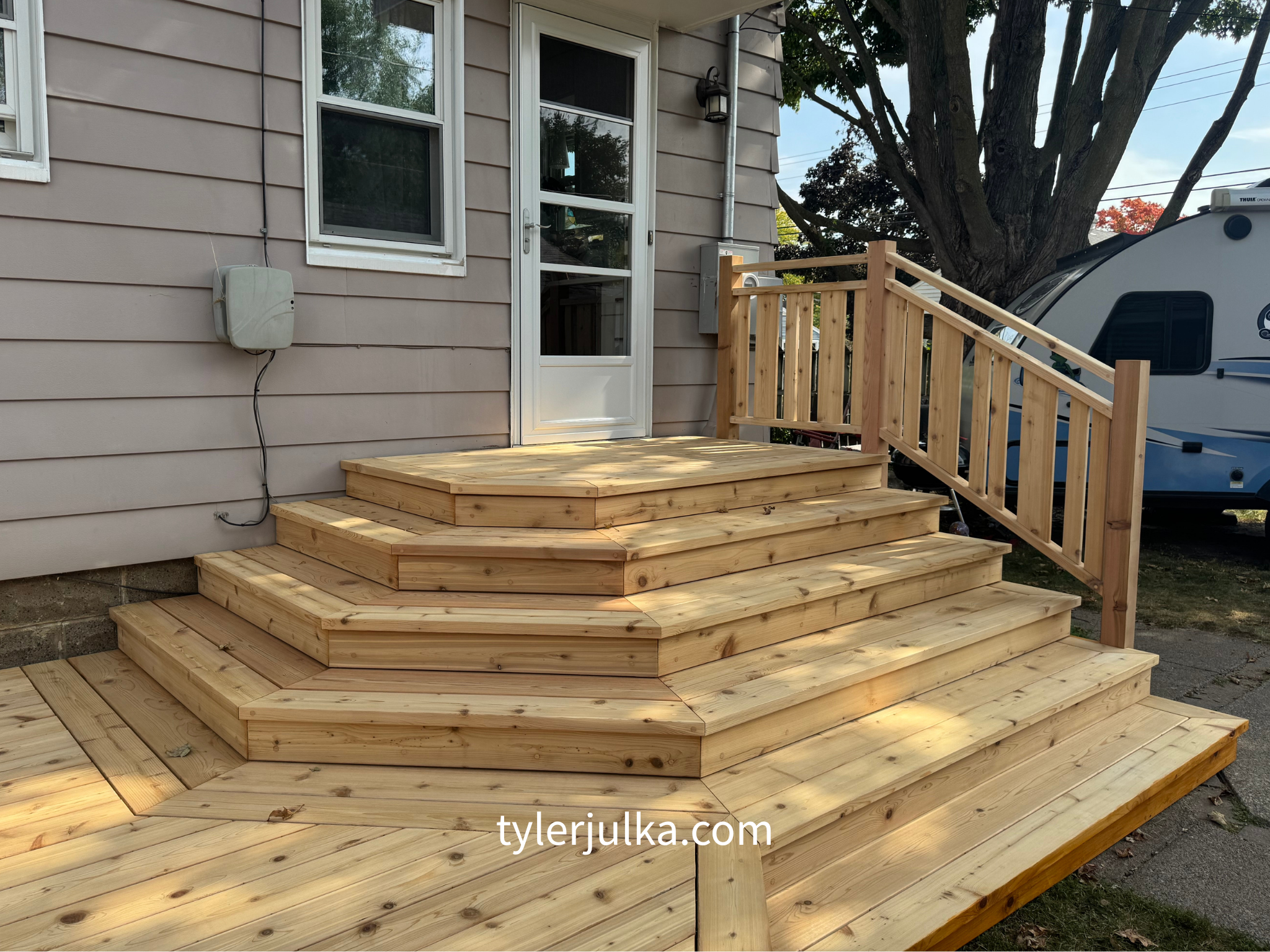 Newly constructed wooden deck with stairs and railing outside a house, with a door, window, and trees in the background.