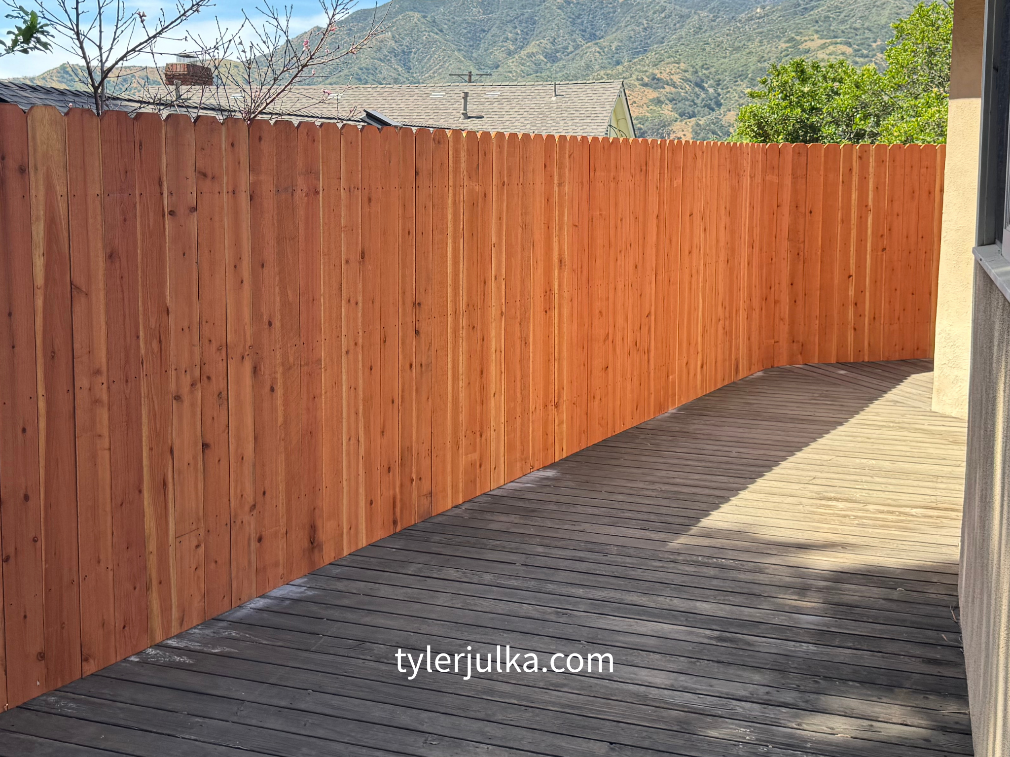 A curved wooden deck with a new wooden fence, mountains in the background, and a house with a rooftop in a suburban setting.