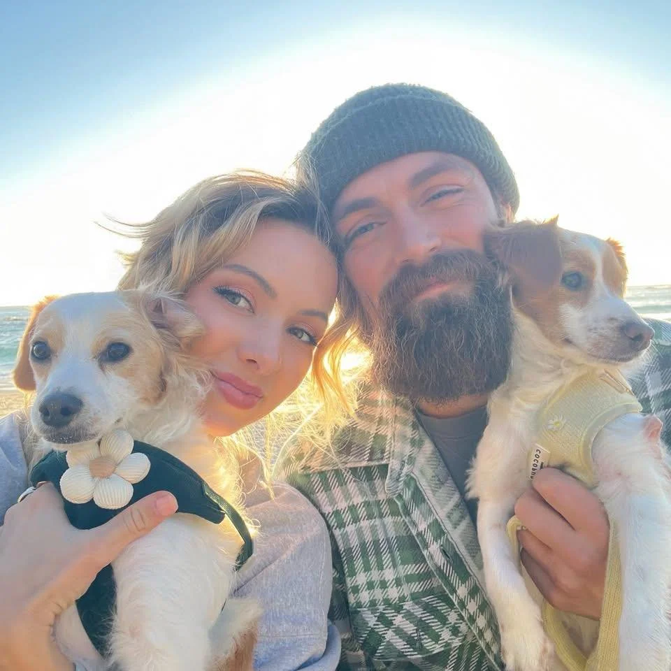 A couple holding two small dogs on a beach at sunset.