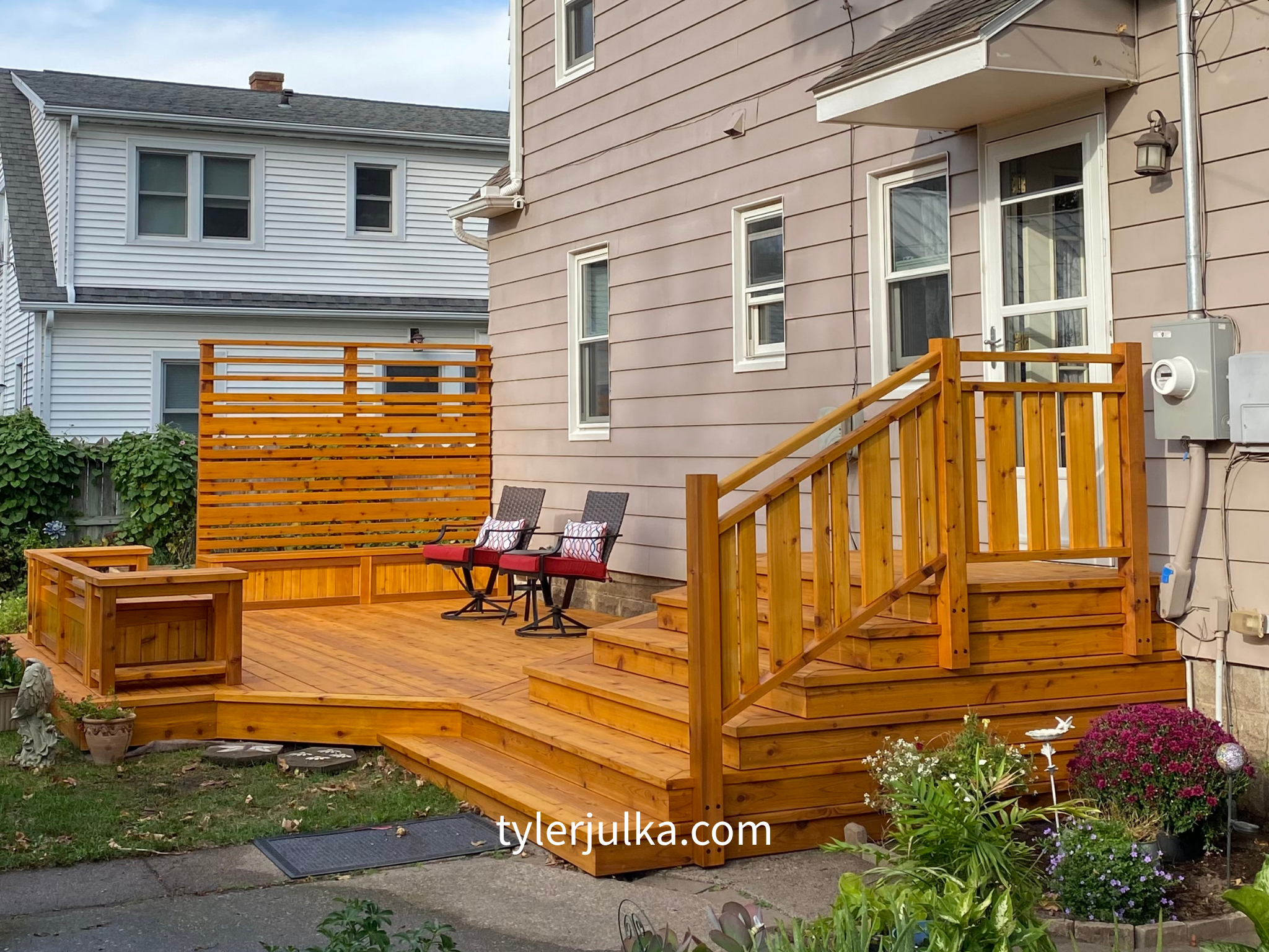 A newly built wooden deck with steps, railing, and outdoor furniture attached to a house with beige siding and several windows. There are chairs with red cushions, a potted plant, and decorative flowers around the deck.