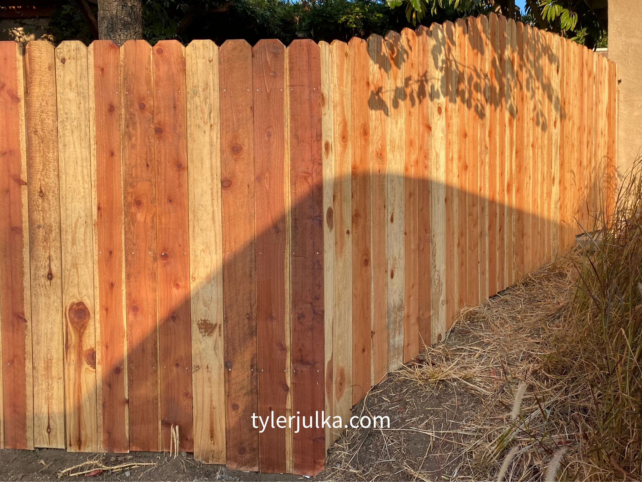 New wooden privacy fence installed outdoors next to dry grass and vegetation with trees in the background.