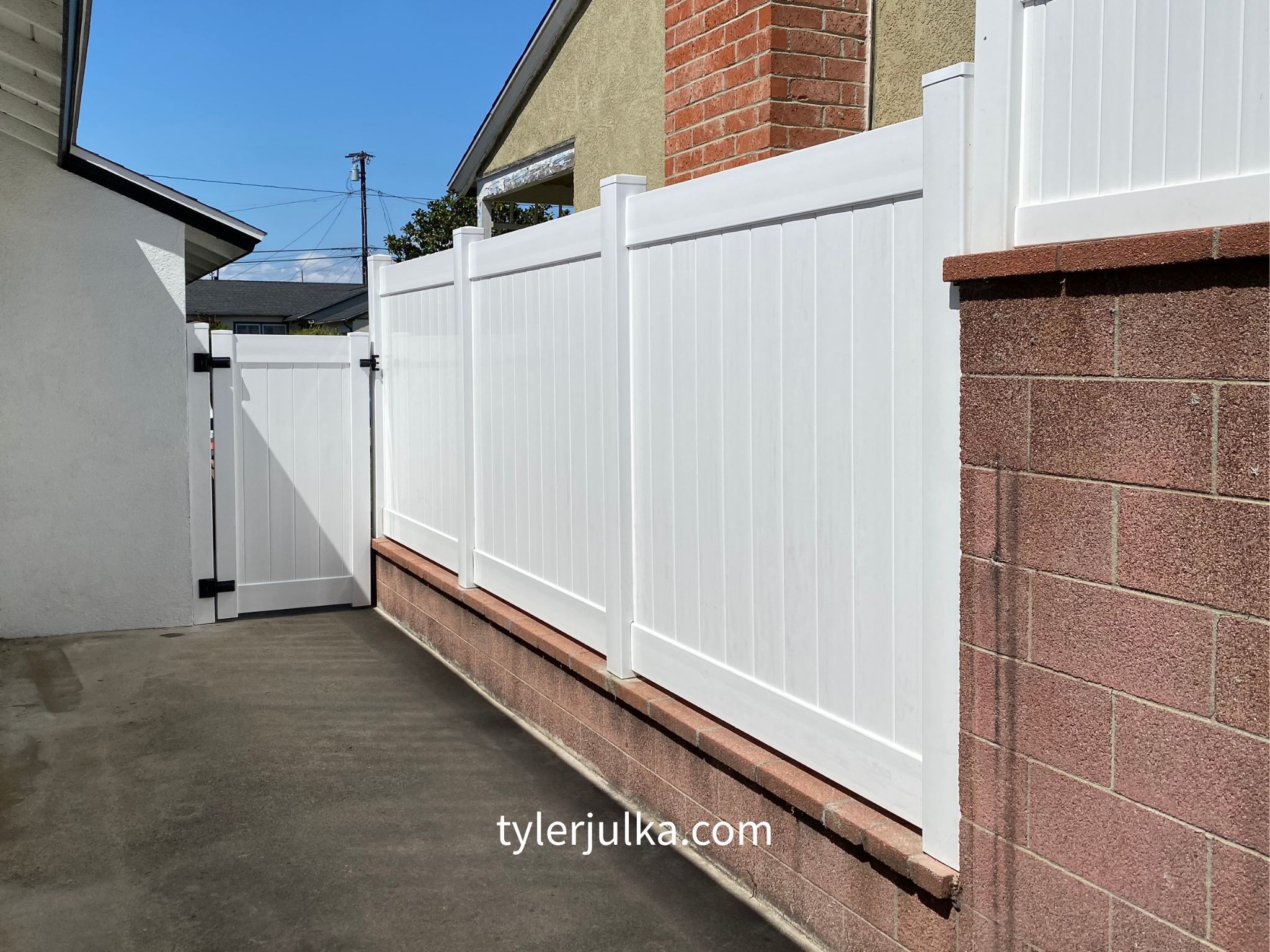 White vinyl privacy fence with a gate, installed on a brick wall base, along a concrete patio, under a clear blue sky.