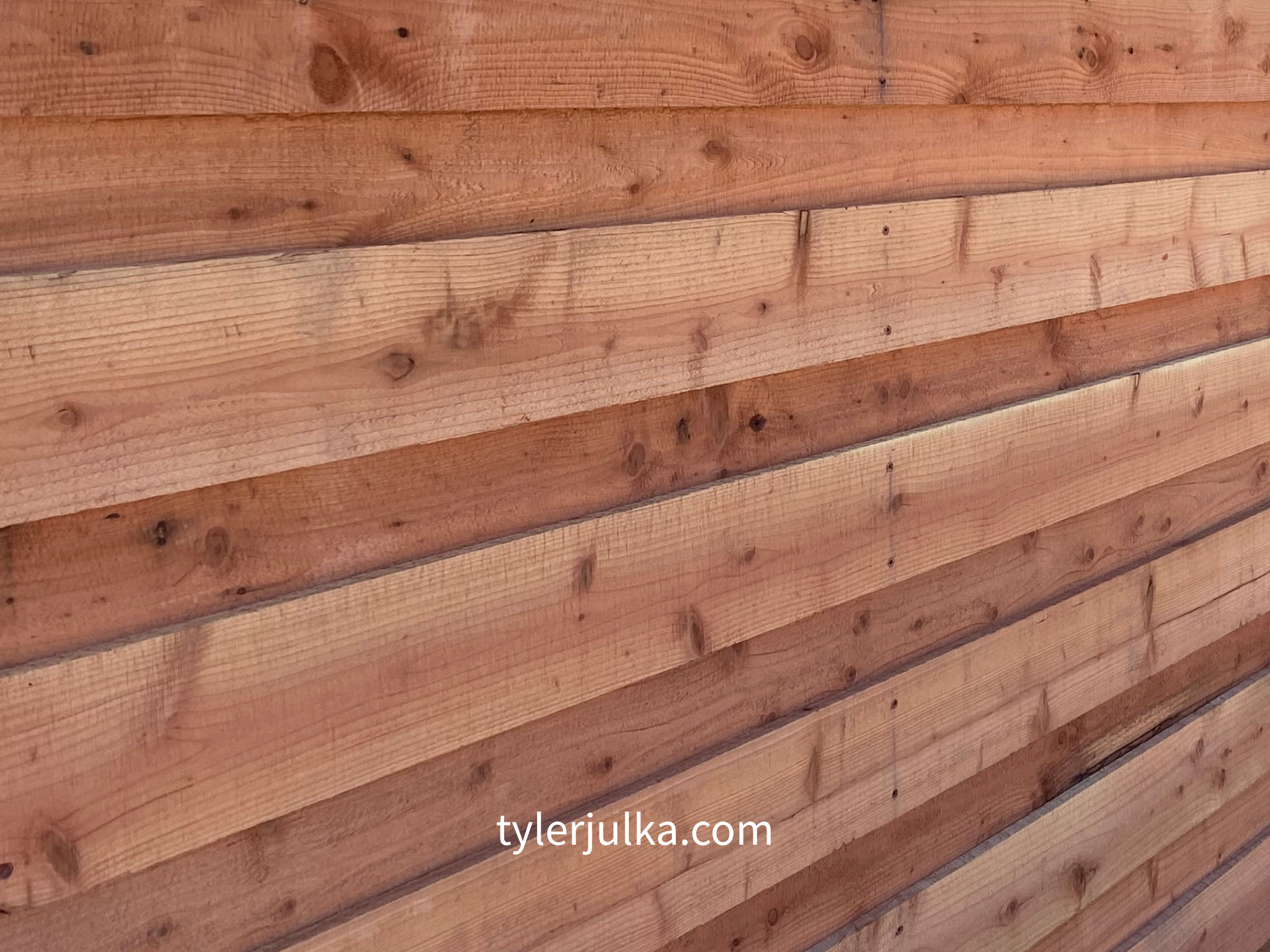 Close-up of a wooden wall made of horizontal planks with natural wood grain and knots, with the website 'tylerjulka.com' written at the bottom.