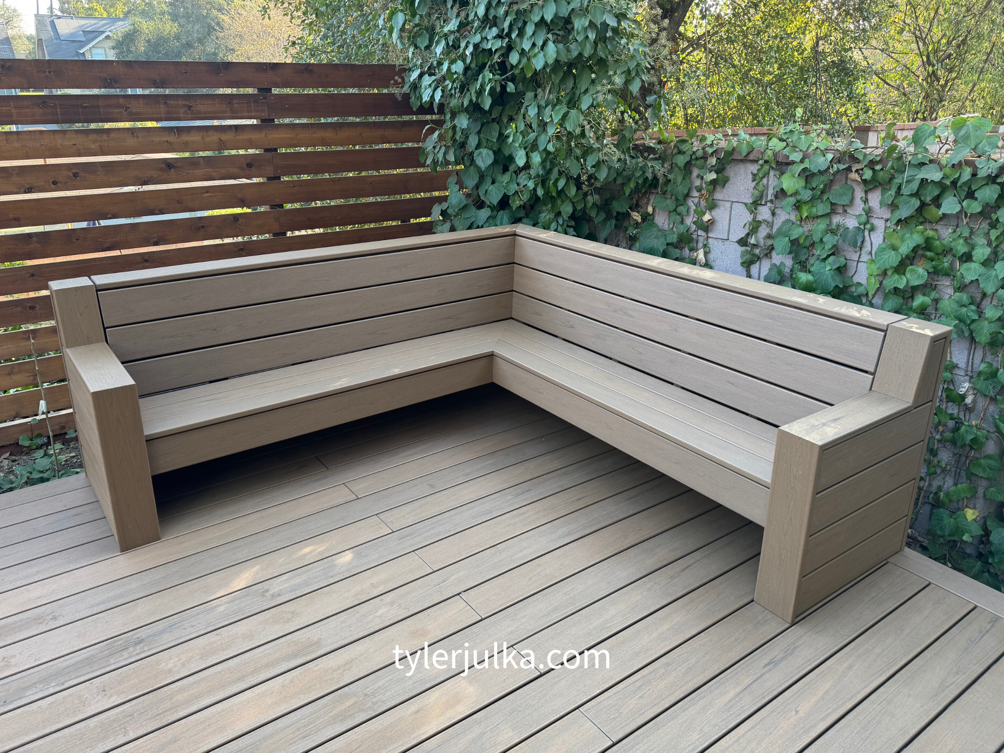 Empty wooden outdoor bench on a deck with horizontal planks, surrounded by a wooden slat fence and a wall covered with green ivy leaves.