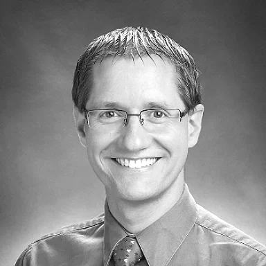A black-and-white portrait of a young woman with long hair smiling at the camera, wearing earrings and a dark top.