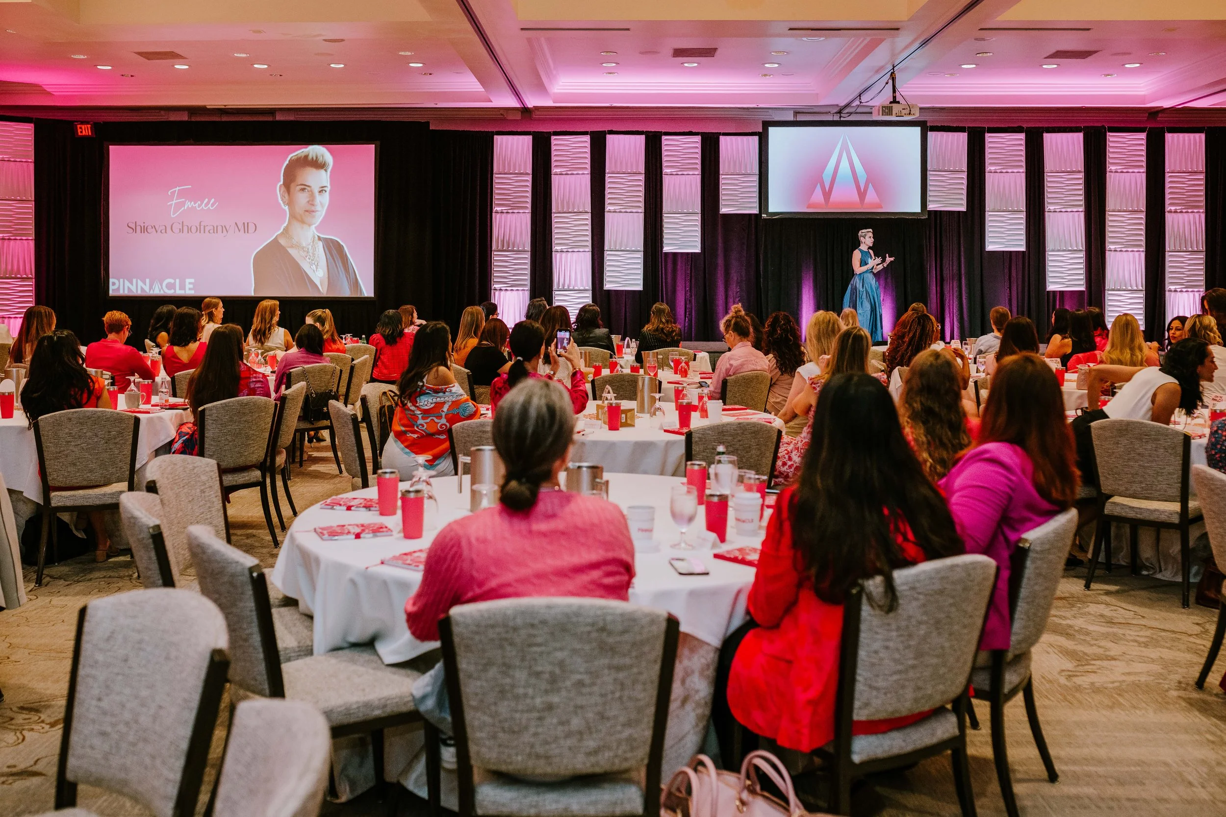 A large conference room filled with women attending a presentation. The stage has two screens, one showing a woman in a blue dress and the other displaying a slide with a woman's photo and the name 'Shieva Ghofrany MD'. The room is decorated with pin
