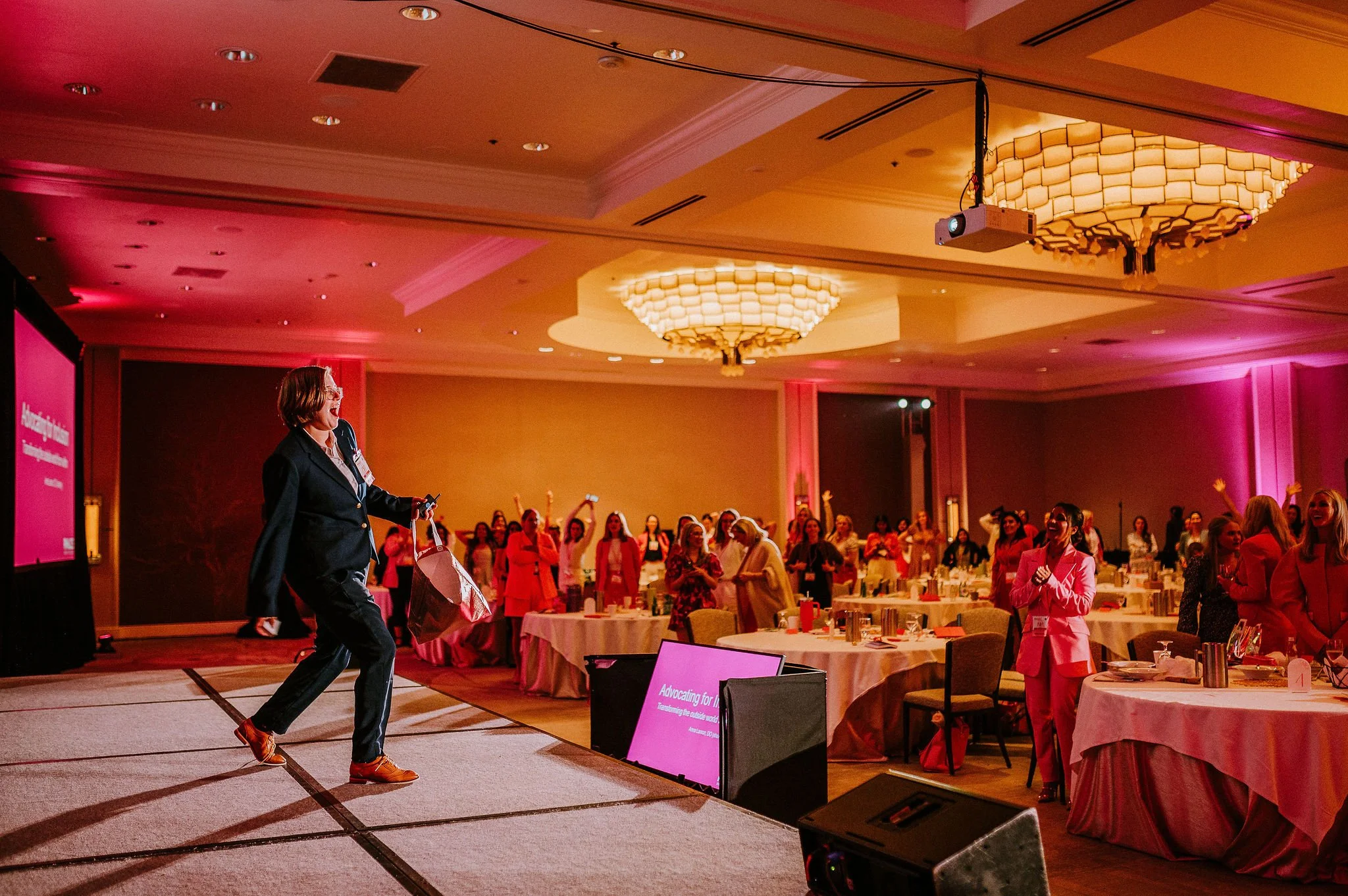 Person on stage at a conference with audience watching, tables with pink lighting.
