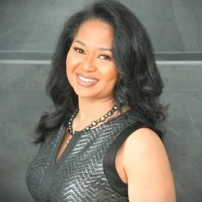 Smiling person with long black hair wearing a dark sleeveless top and necklace in front of a gray background.