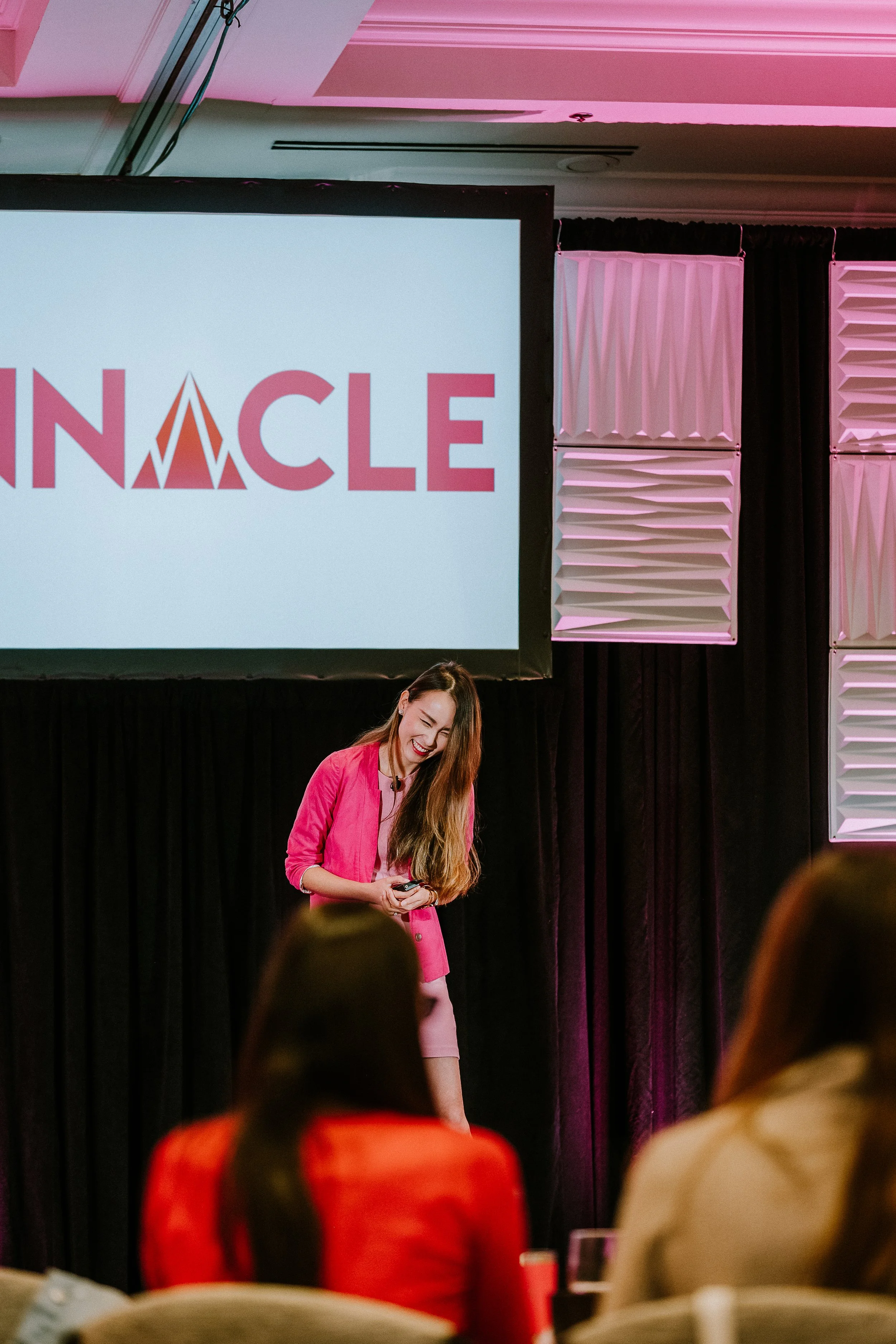 A woman in a pink blazer standing on stage, smiling and looking down, during a presentation or speech at a conference or event. Behind her is a large screen displaying part of a logo with the word 'INNACLE' and pink and white decorations.