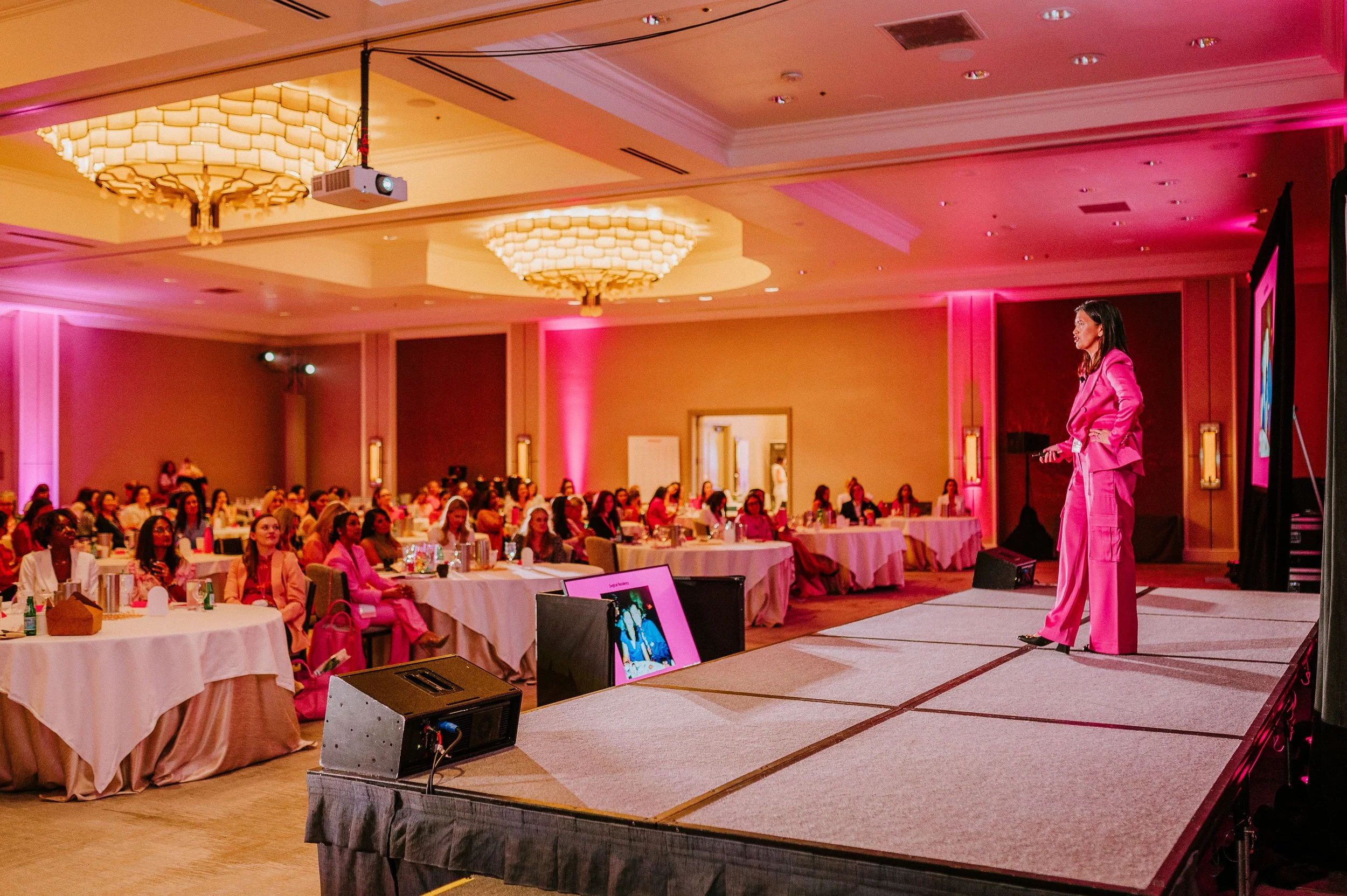 A woman in a pink suit speaks on a stage at a conference, addressing an audience seated at round tables in a pink-lit room with chandeliers.