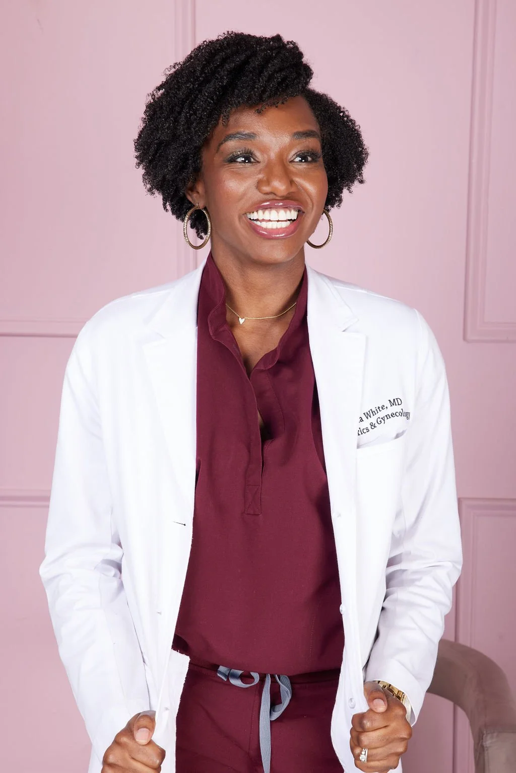 Smiling person in a white coat standing against a pink background.