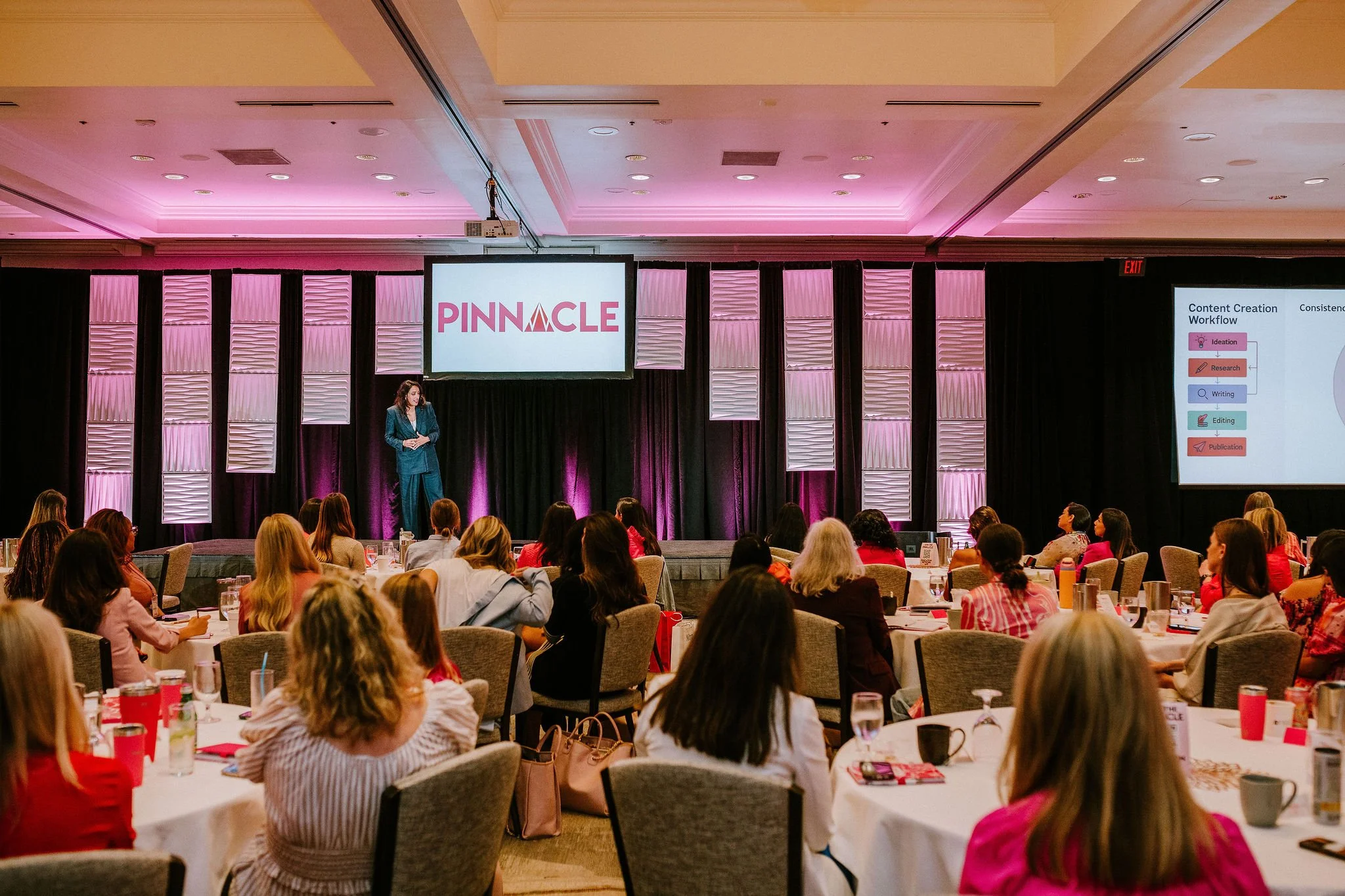 A woman speaker on stage at a conference, presenting to an audience of women. The stage has a backdrop with pink and white panels, and a large screen displays the logo 'PINNACLE' and a slide about content creation workflow. The audience sits at round tables with glasses, notebooks, and coffee mugs.