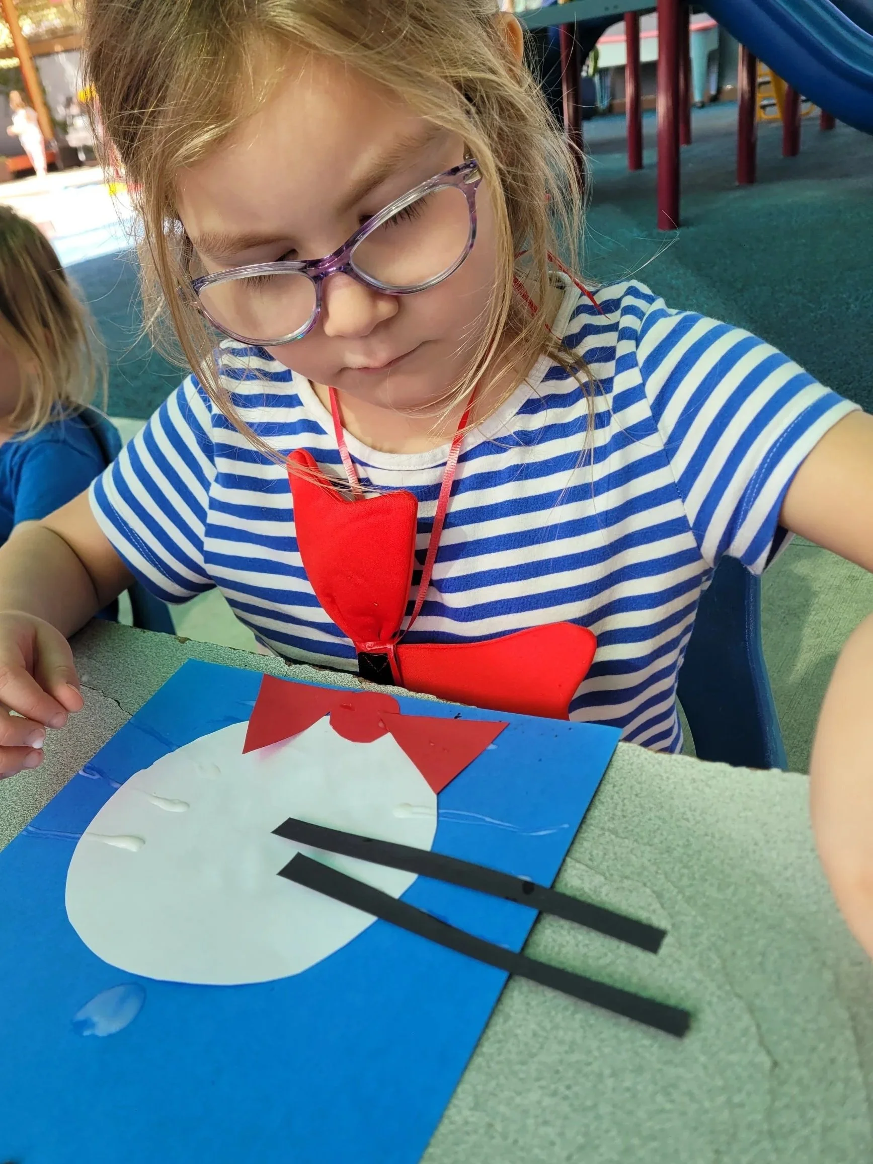 A young girl with glasses and a striped blue and white shirt is looking down, working on a craft project with red and black paper on a blue and white paper fan at a table in a play area.