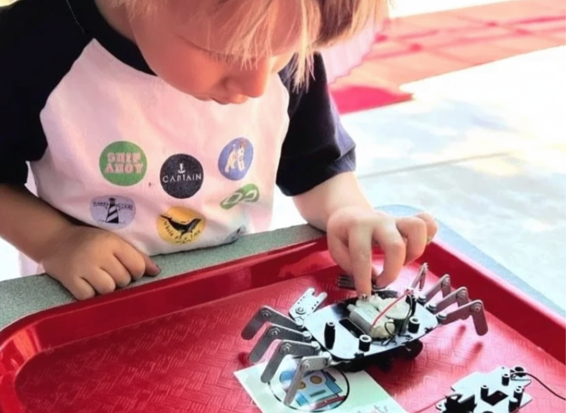 A young boy looking down and building a small robotic crab on a red tray with mechanical parts and wires.