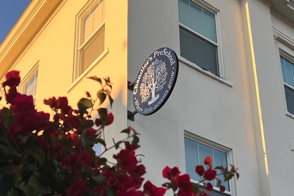 Sign for Chandler's Preschool with a tree logo and slogan that says 'Where little acorns grow' on the exterior of a large white building, with pink flowers in the foreground during sunset.