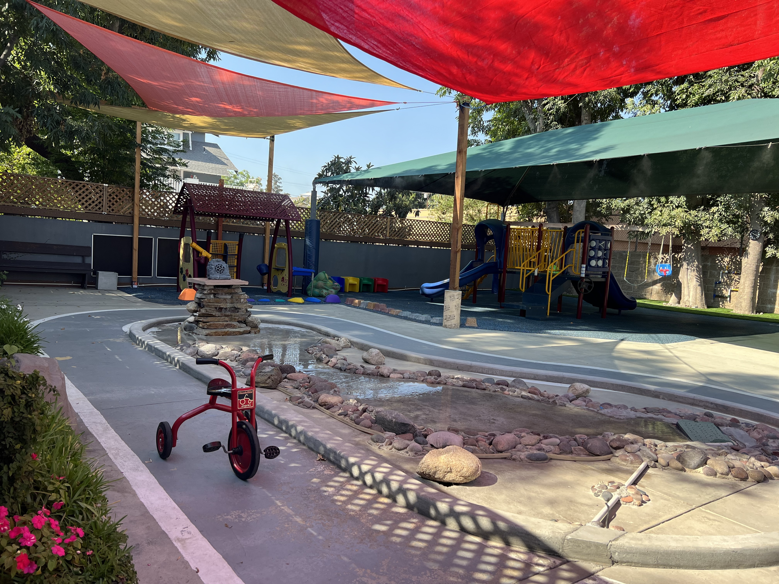 Children's playground area with colorful shade sails overhead, a small fountain made of rocks, playground equipment including slides, and a concrete pathway with a red ride-on toy and pink flowers nearby.