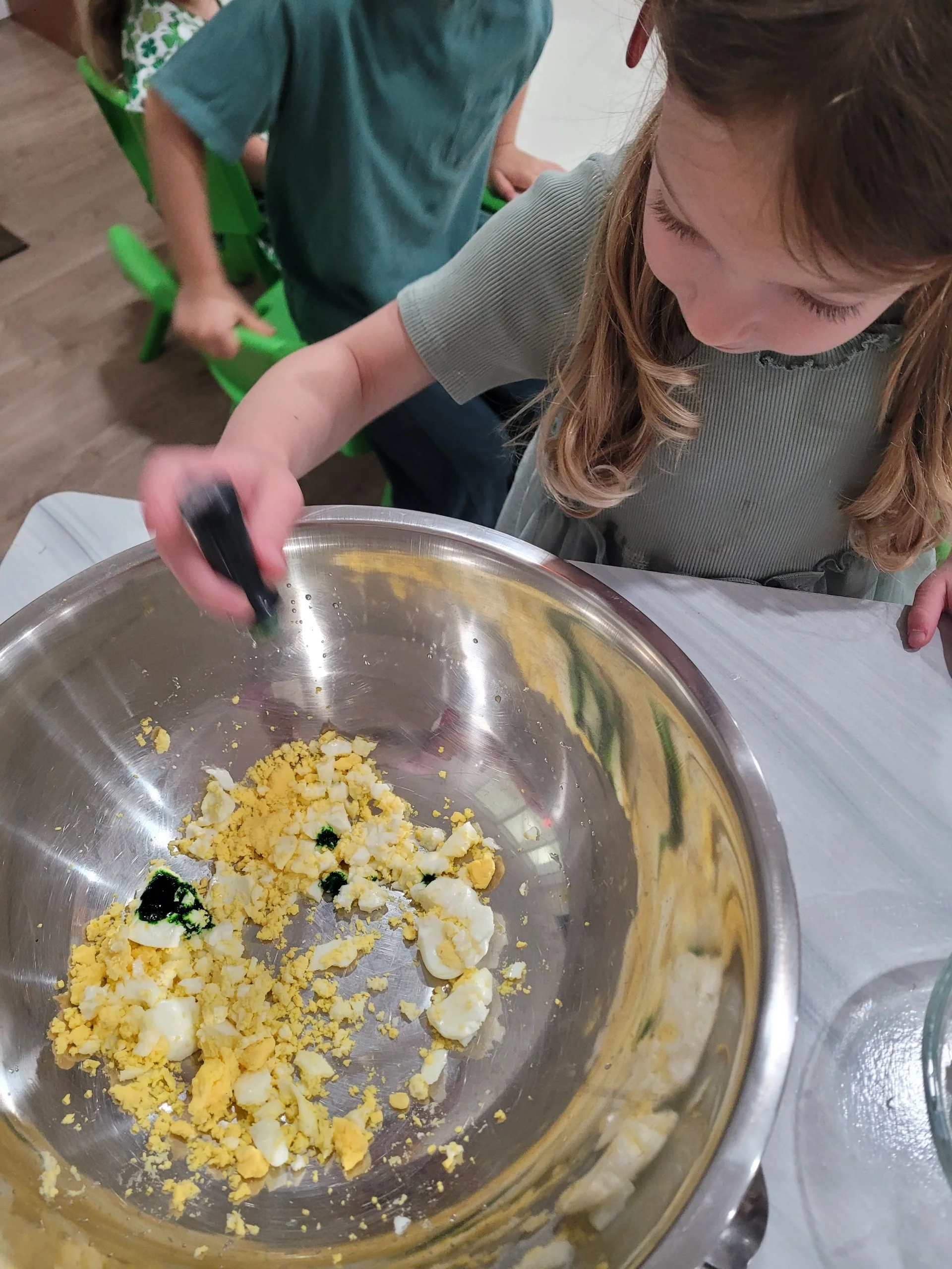 Close-up of a young girl with long hair, wearing a gray shirt, mixing chopped hard-boiled eggs with green food coloring in a stainless steel bowl.