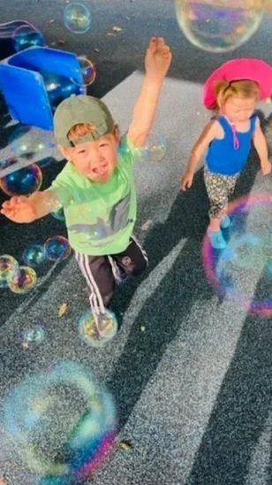Two young children playing outdoors with soap bubbles, one boy excited with his arms raised and a girl wearing a pink hat and backpack.