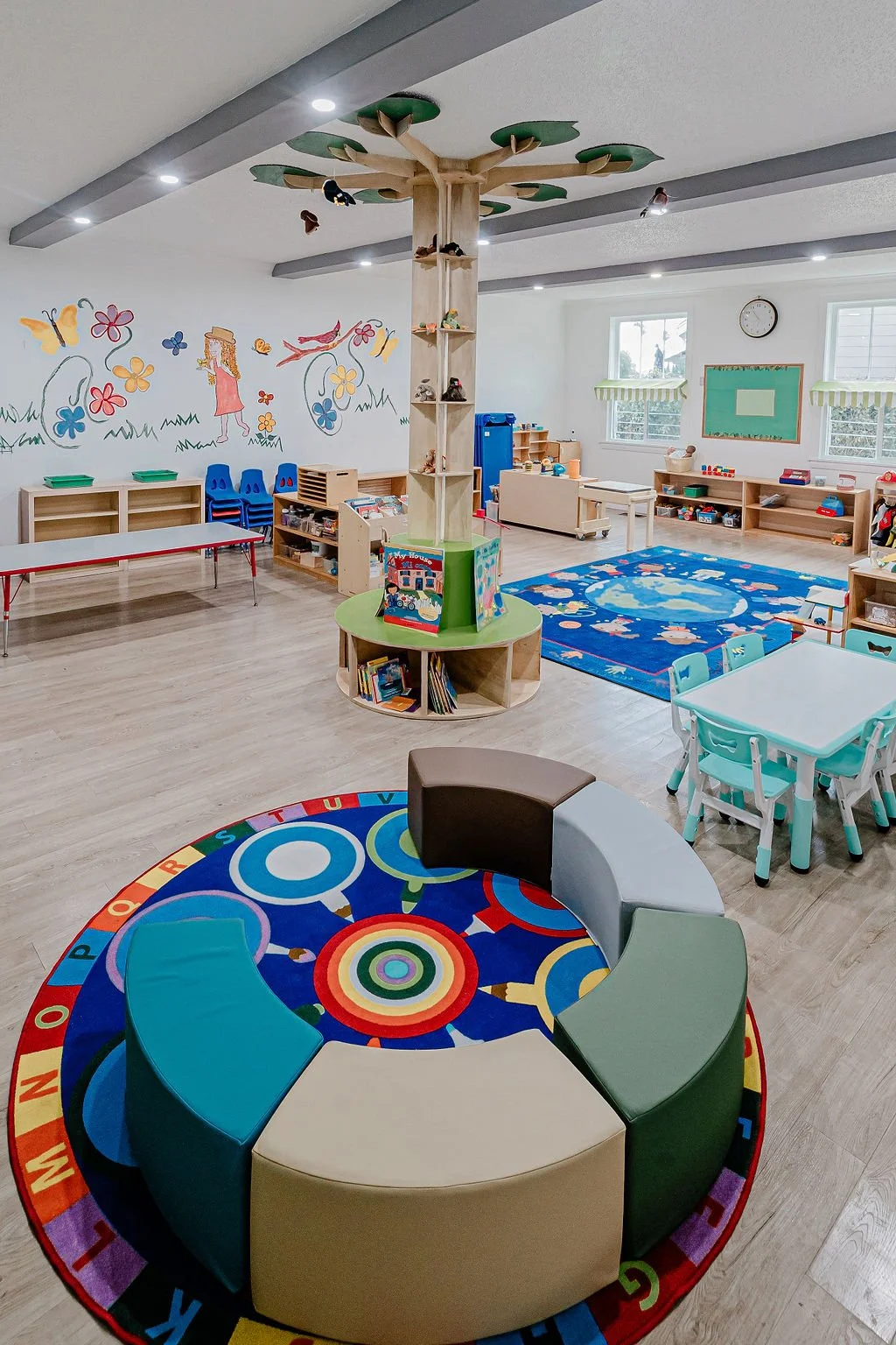 A colorful and organized preschool classroom with a circular seating area on an alphabet-themed rug, a play tree with shelves, tables with chairs, a blue carpet with animal illustrations, and educational toys on shelves. The room is bright with natural light and decorated with murals of flowers, butterflies, and a girl on the wall.