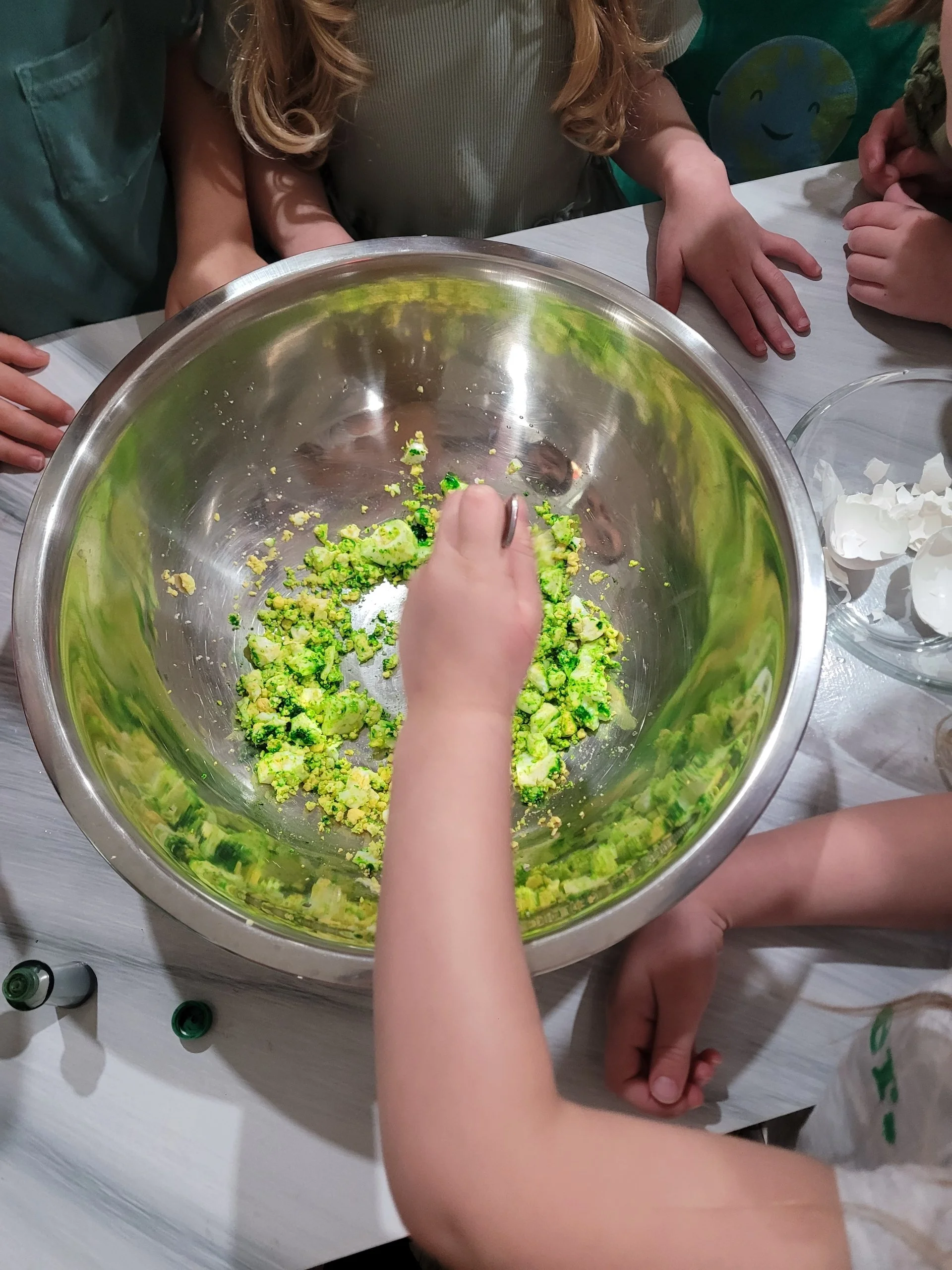 Children gathered around a table watching as one of them mixes or crushes green food coloring into lighter colored food in a stainless steel bowl.
