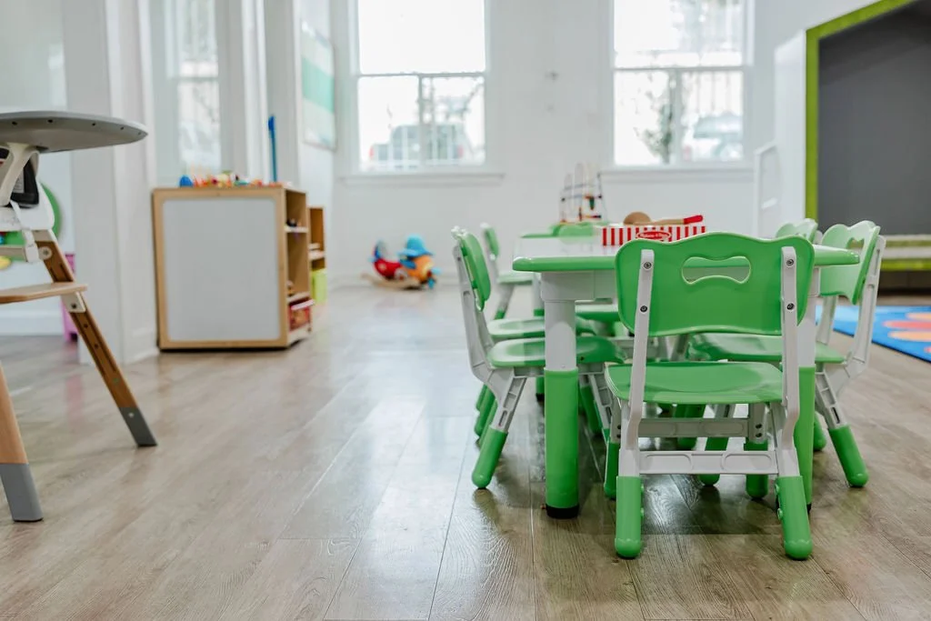 A brightly lit children's playroom featuring a small table with green and white chairs, a chalkboard, a bookshelf with toys, and a window with a view outside.