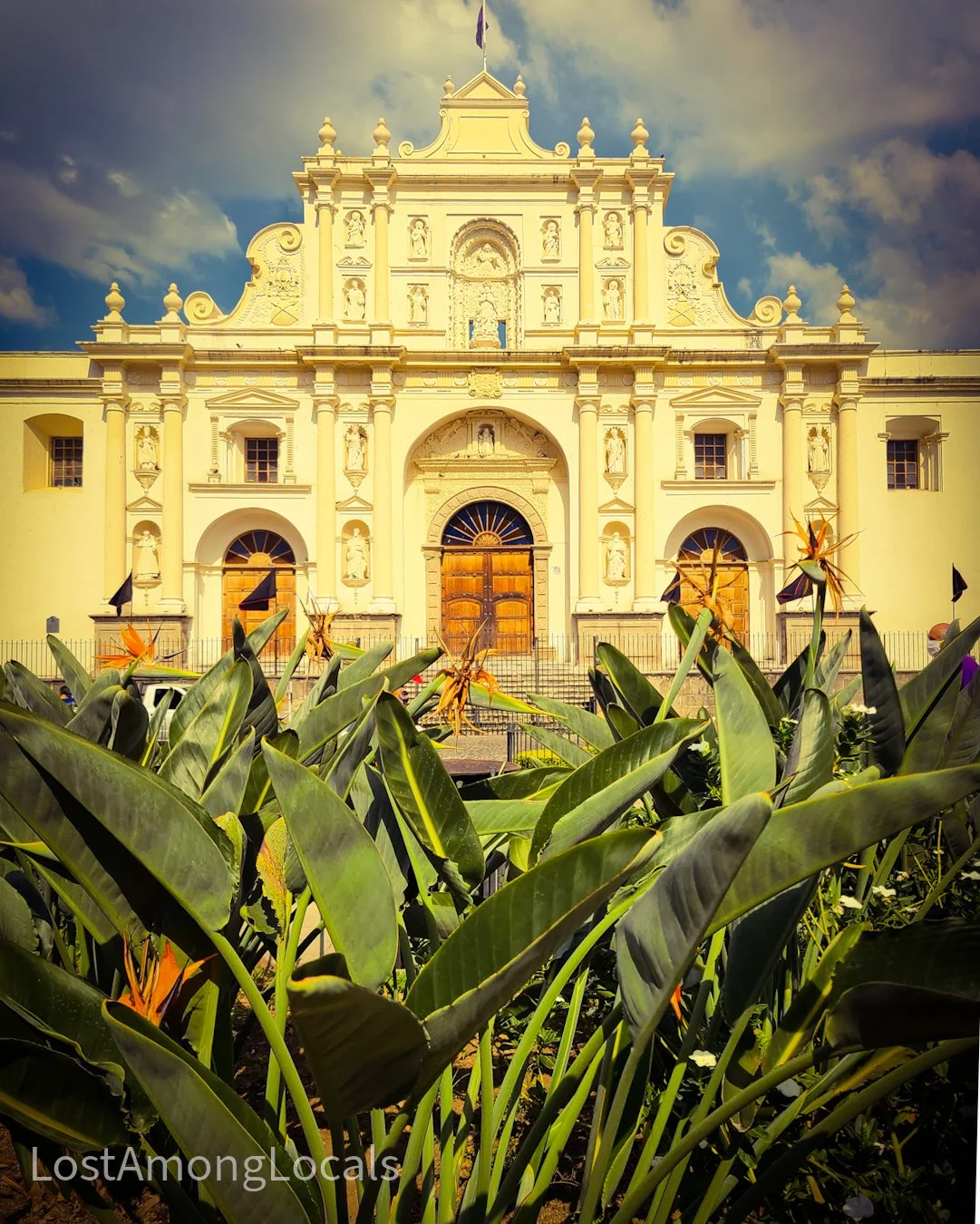 A historic church with detailed white facade behind green plants with orange flowers, where the sky is partly cloudy.