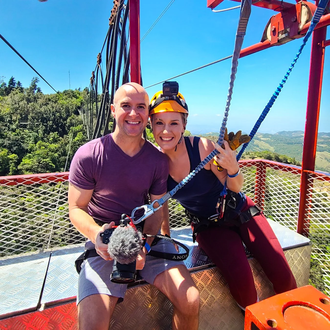 A man and a woman sitting together on a metal platform at the top of a tall structure, both smiling at the camera. The woman is wearing a helmet with a camera, holding ropes, and has climbing gear. The background shows green hills and blue sky.