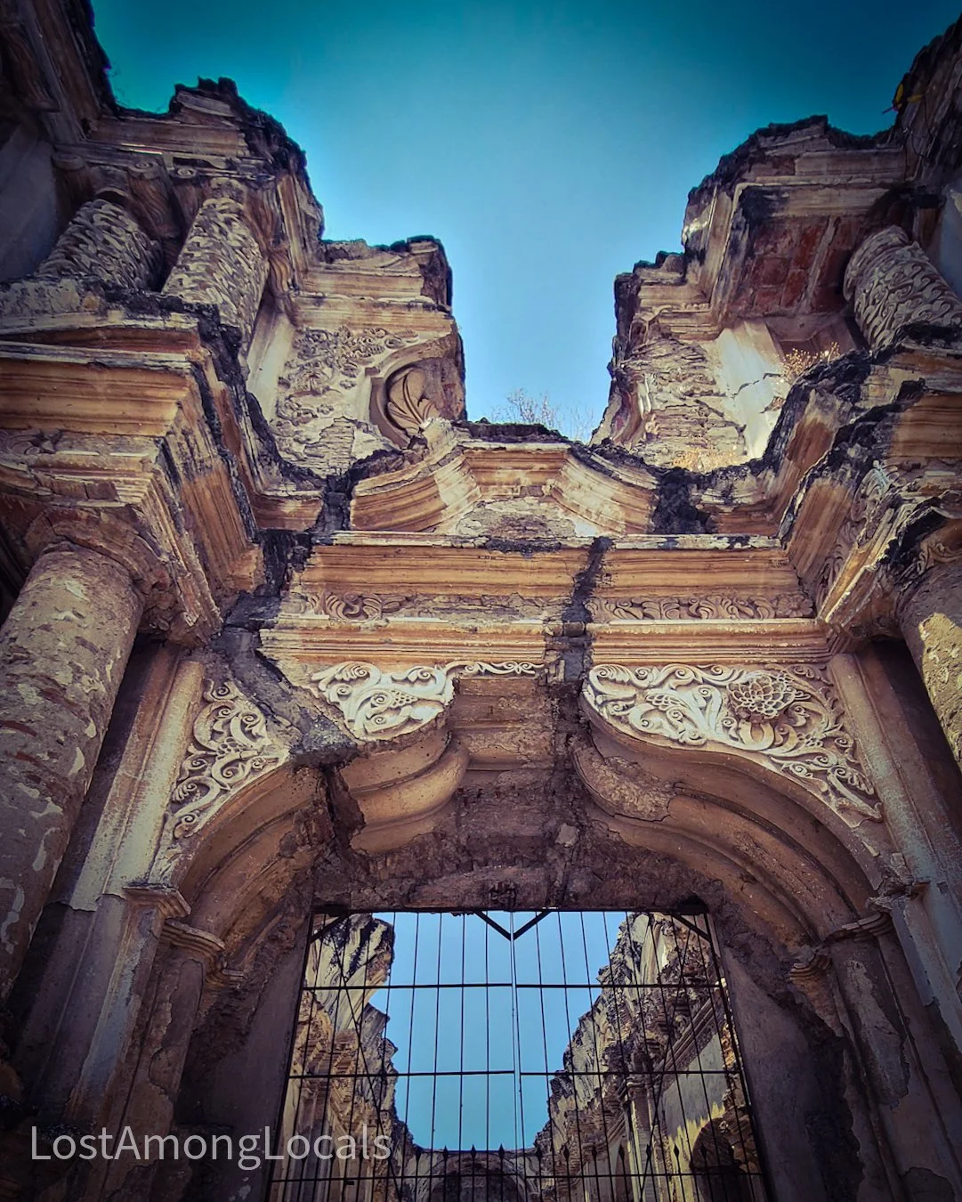 Ruined ancient structure with detailed carvings and columns reaching up to the sky, viewed from below through a barred entrance.