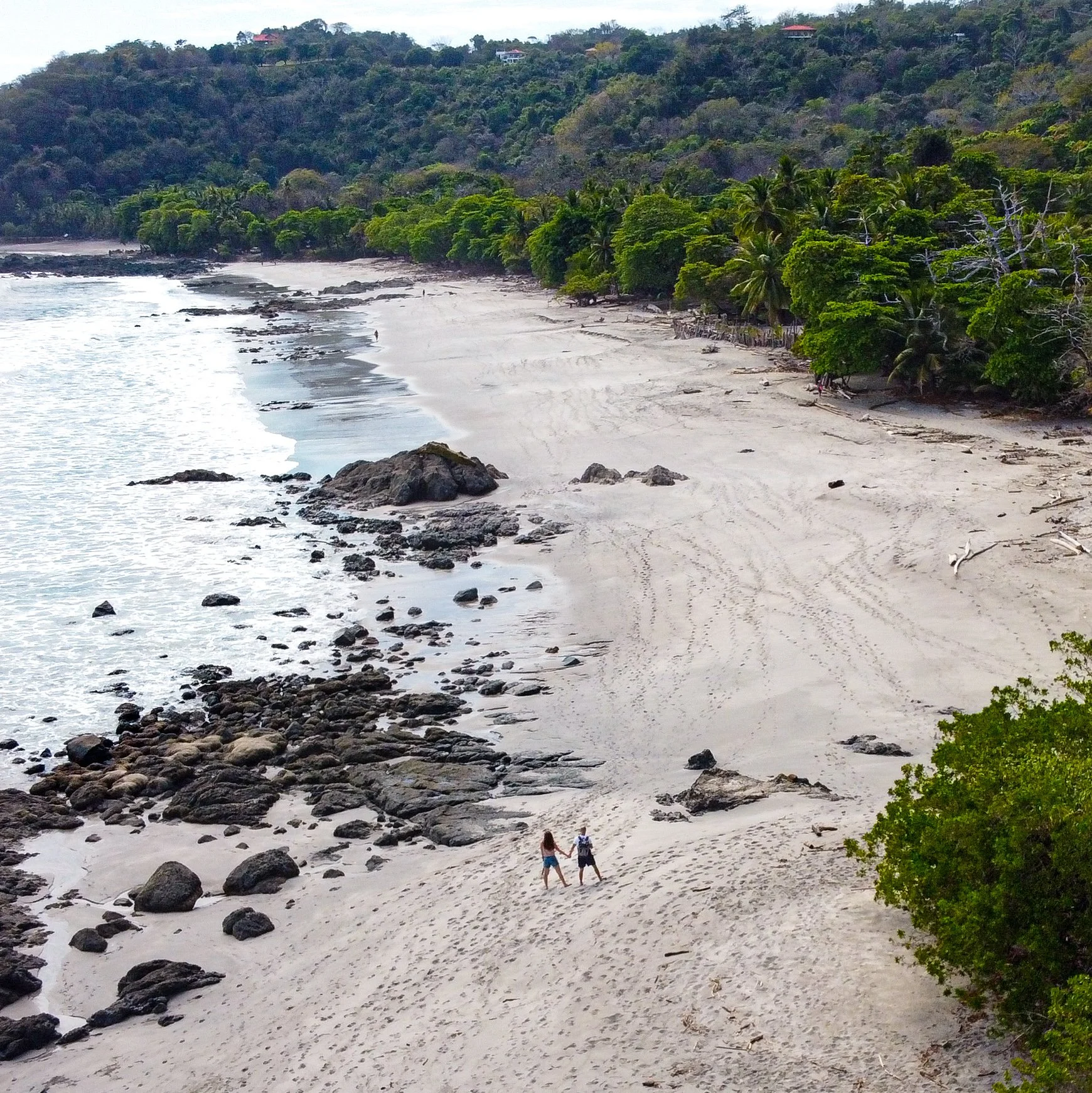 Two people holding hands walking along a sandy beach with rocks, lush green trees, and hillside houses in the background.