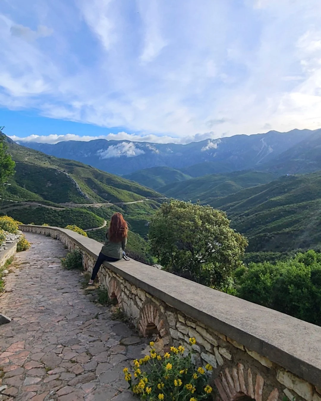 A person with long red hair sitting on a stone wall on a mountain trail, overlooking green rolling hills and distant mountains under a partly cloudy sky.
