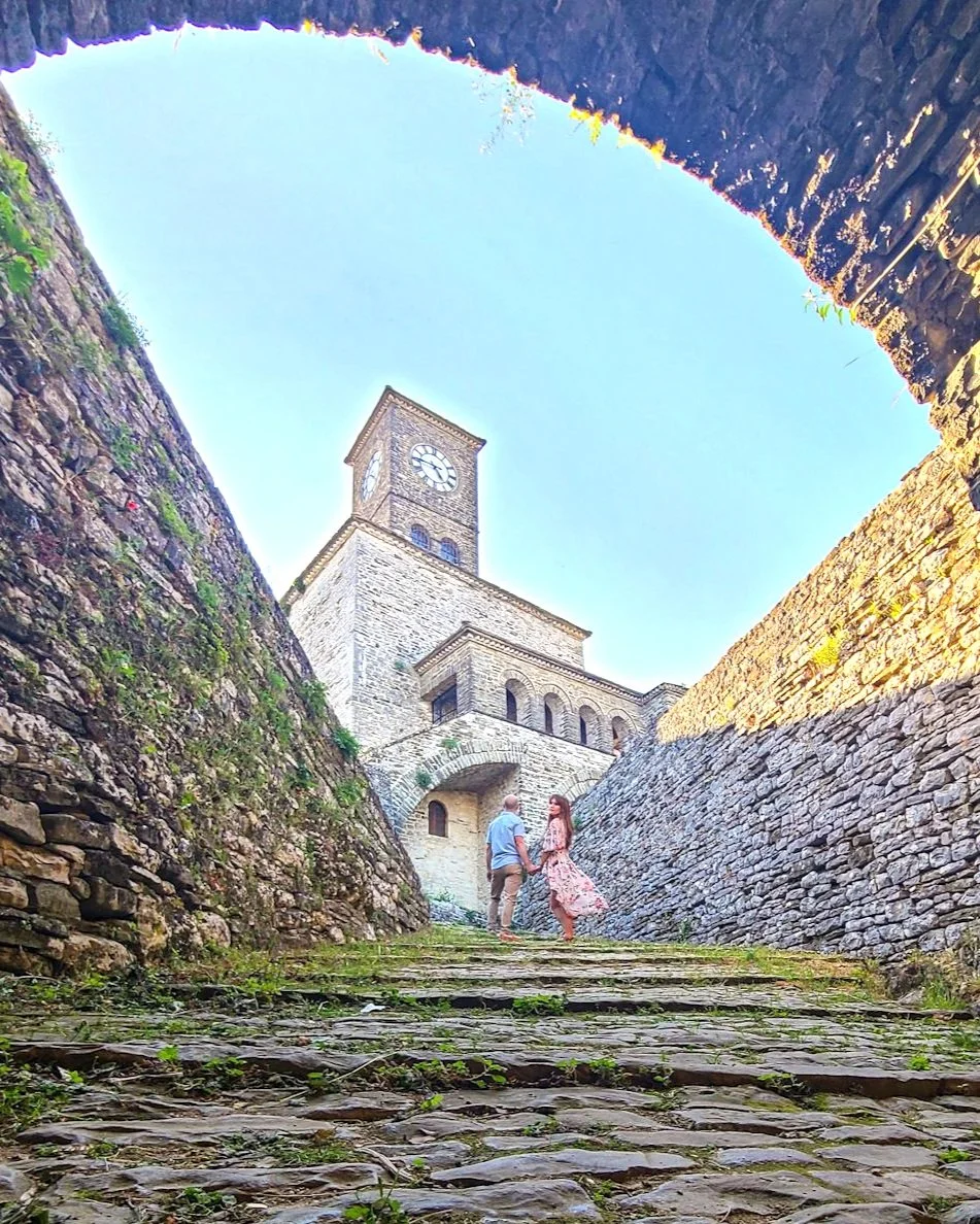 A couple holding hands and walking up a cobblestone path towards a stone clock tower, with tall stone walls on either side, under a clear blue sky.