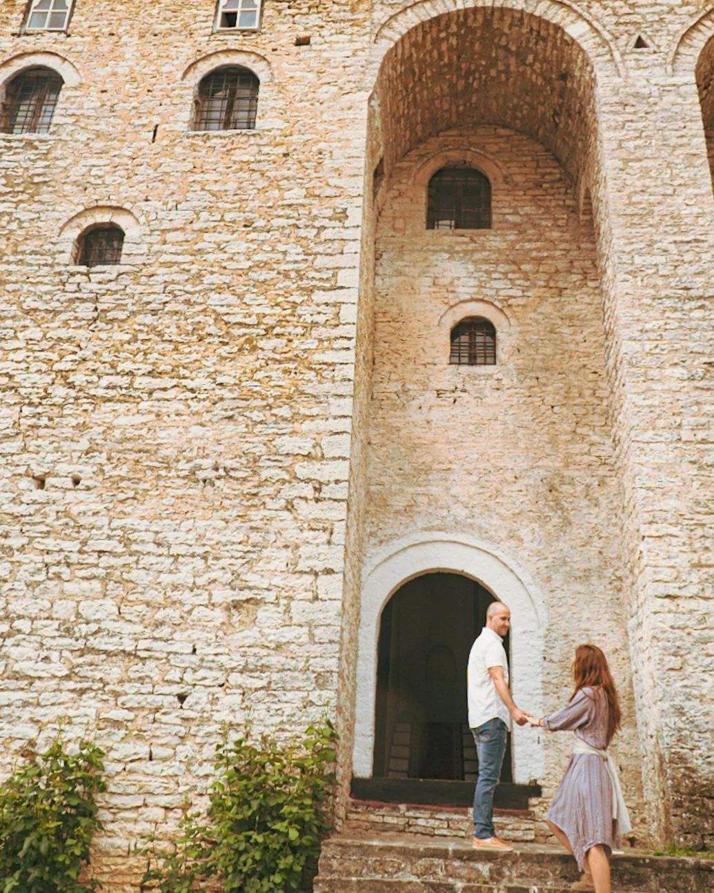 A man and woman holding hands outside a large stone castle with an arched doorway, small windows, and ivy at the bottom.
