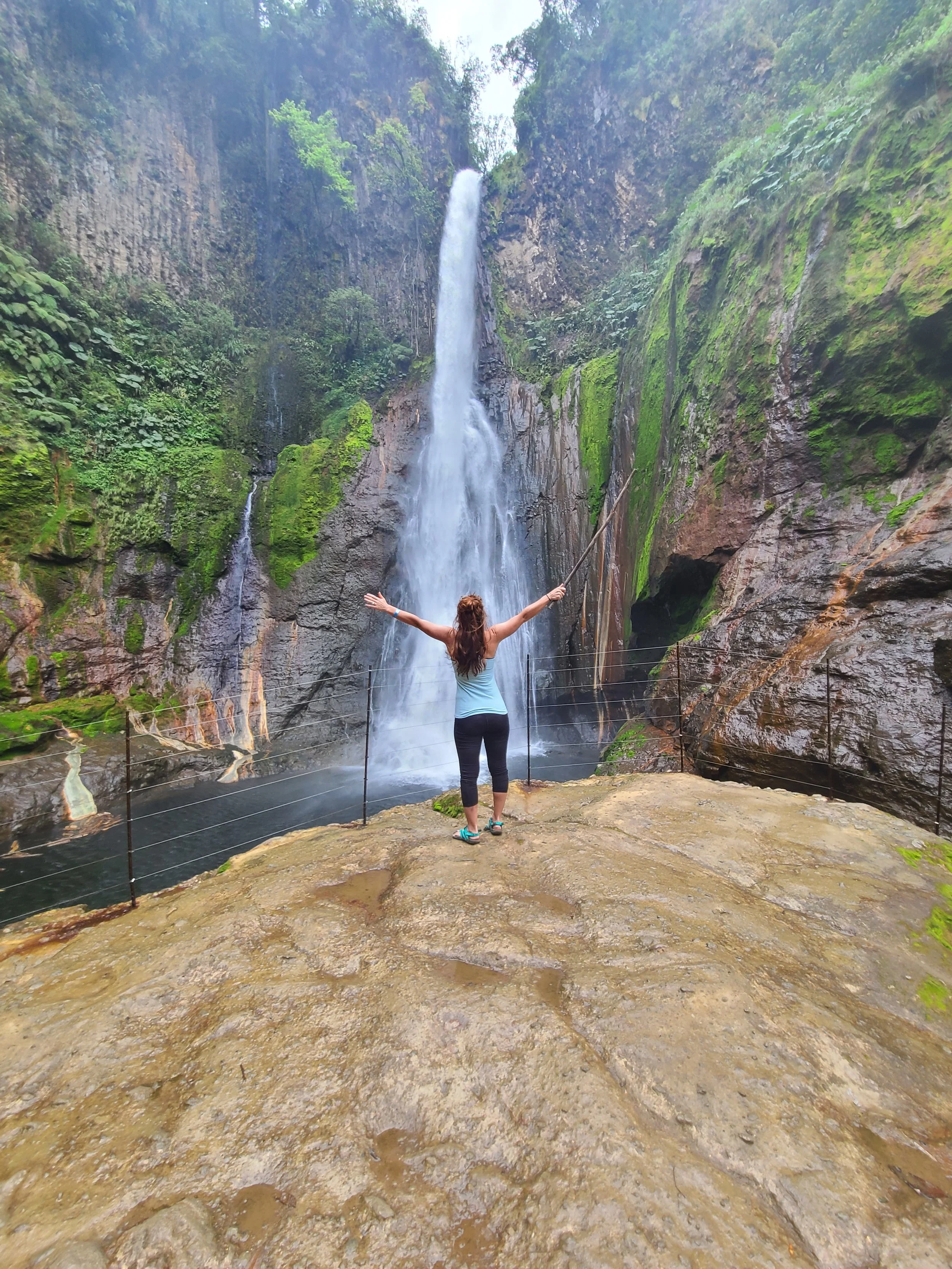 Woman standing with arms outstretched in front of a waterfall in a lush, green, rocky canyon.