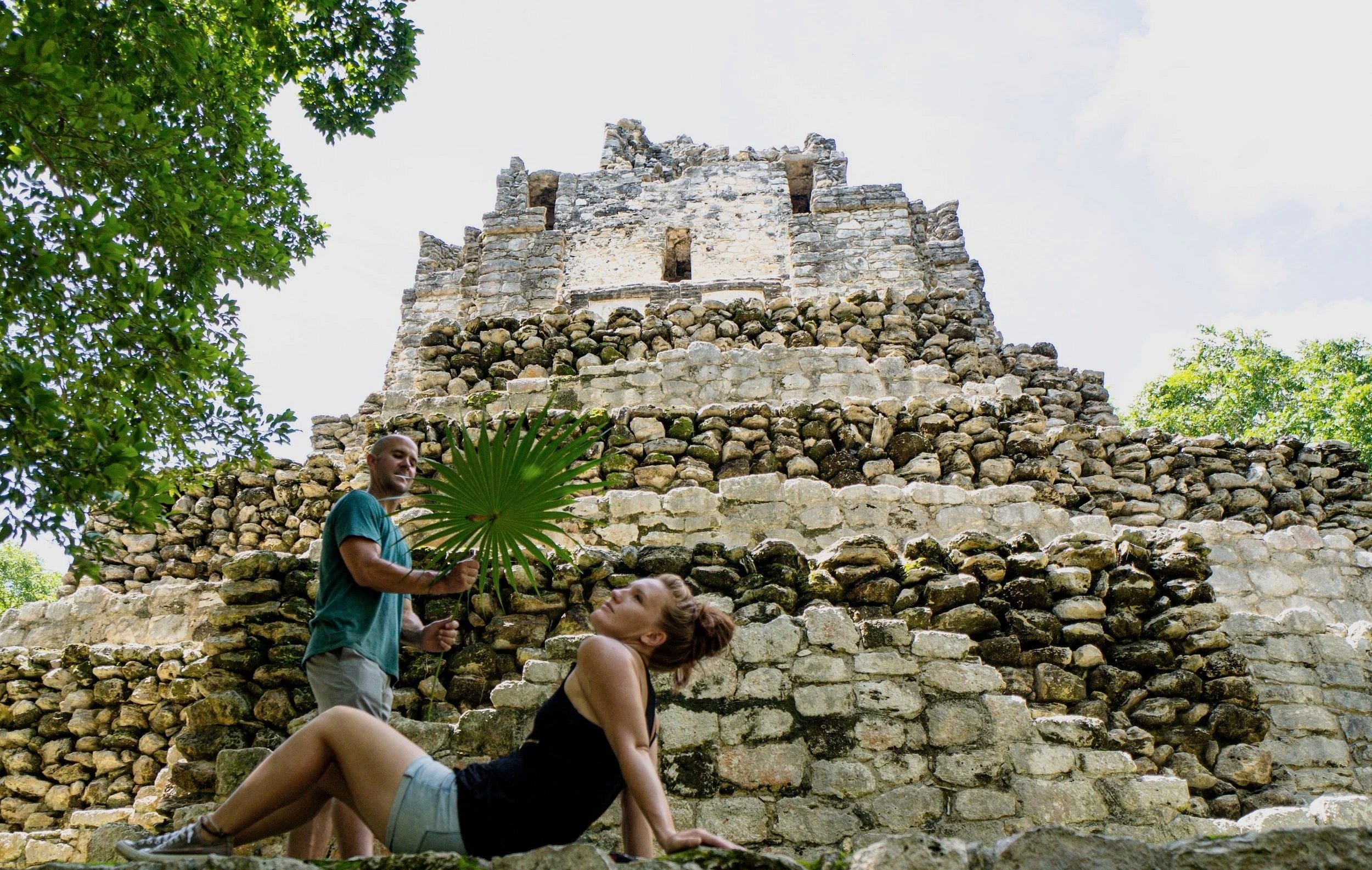 Two women and a man posed on the steps of an ancient stone pyramid with greenery around, one woman reclines on the steps while the man holds a large leaf, and the other woman looks up at him.