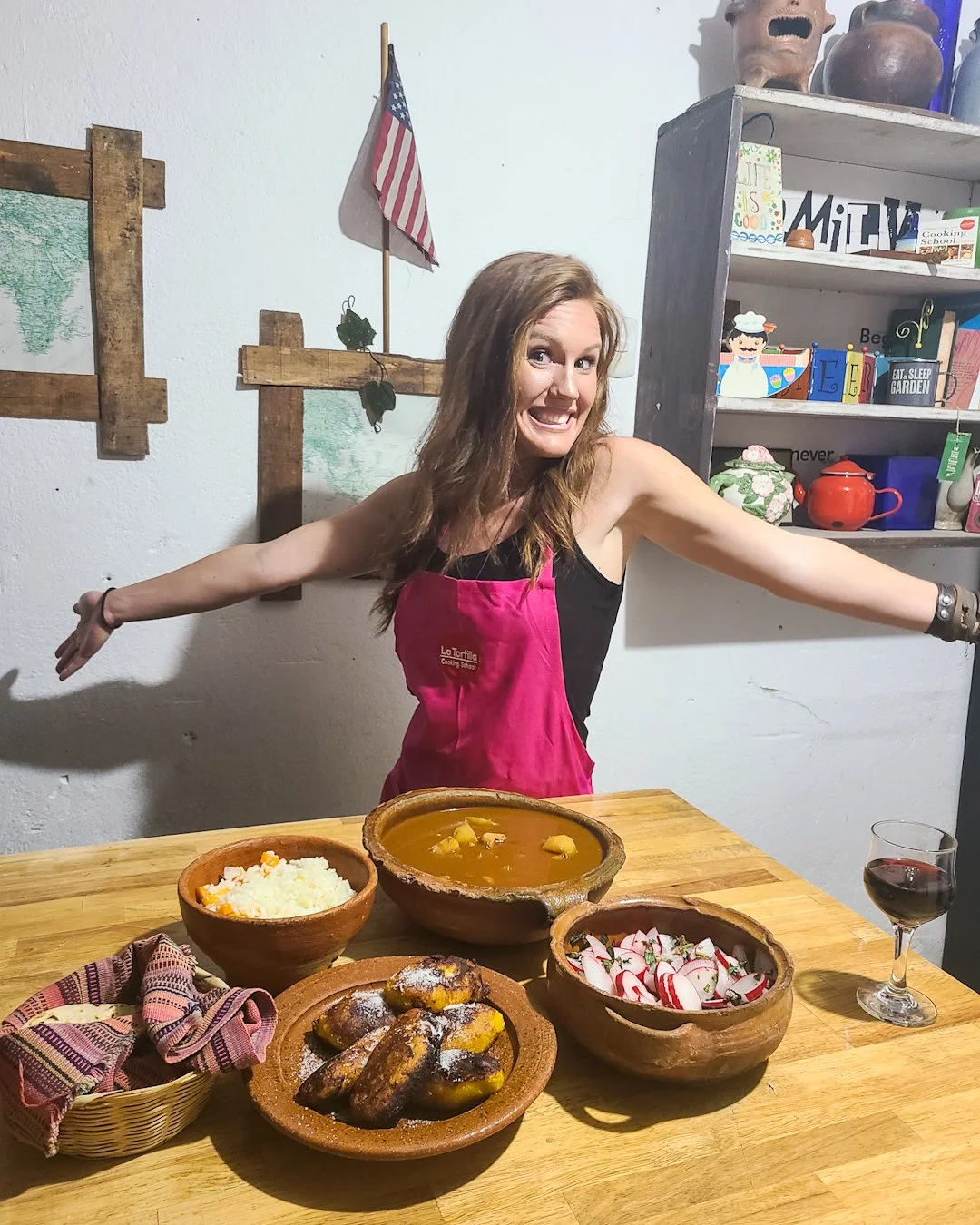 A smiling woman wearing a pink apron stands behind a wooden table with Mexican food dishes, including rice, tamales, a bowl of stew, and a salad. She has arms outstretched and is in a cozy kitchen decorated with American and Mexican-themed items.