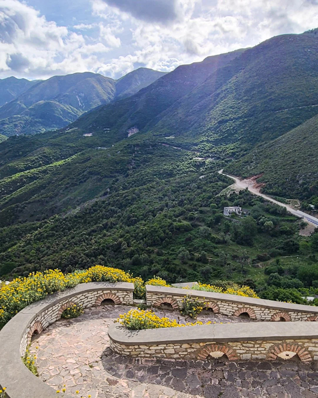 Stone balcony with yellow flowers overlooking lush green mountains and a winding road.