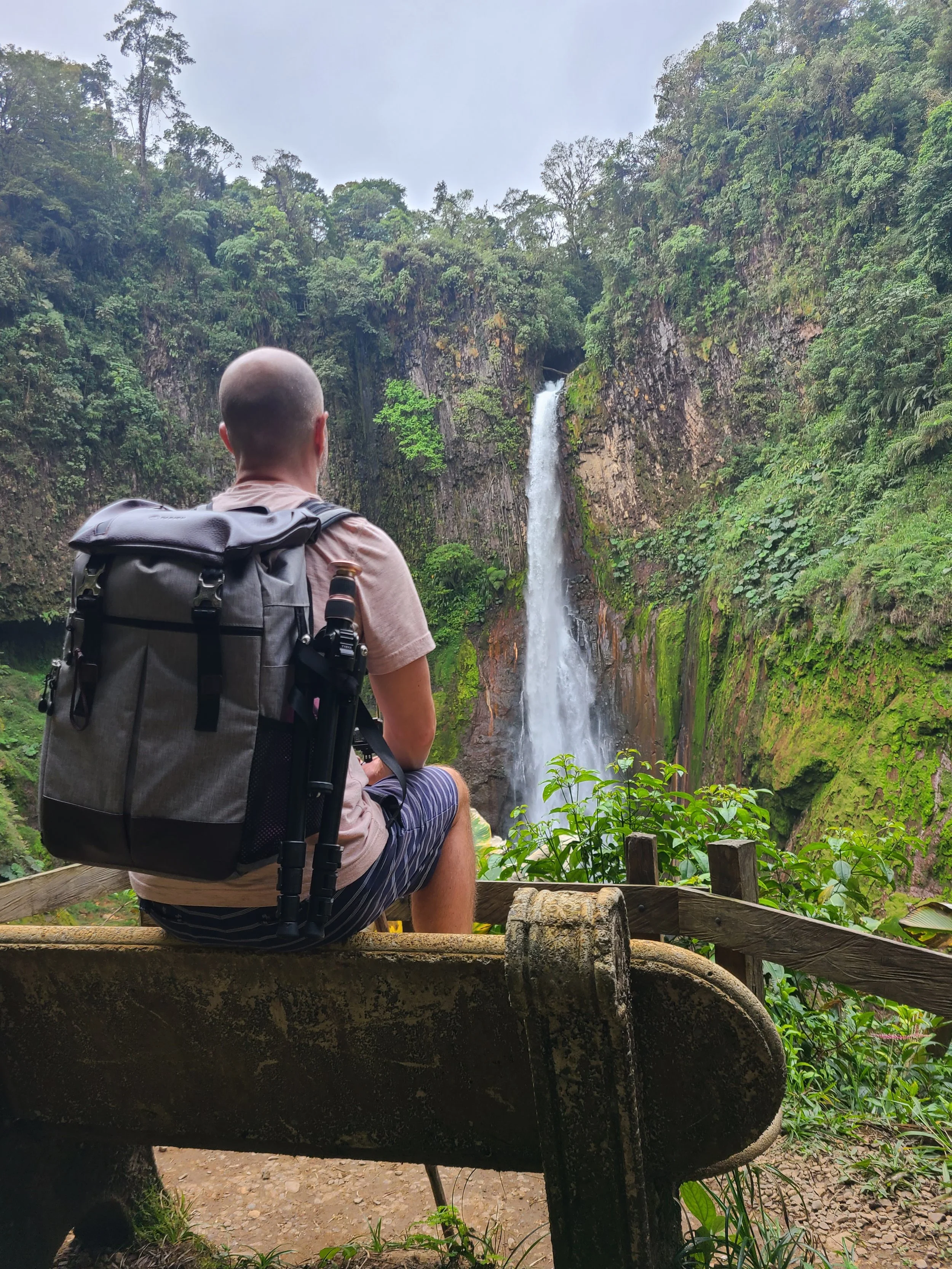 A person with a backpack sitting on a stone park bench, looking at a waterfall in a lush green forest.