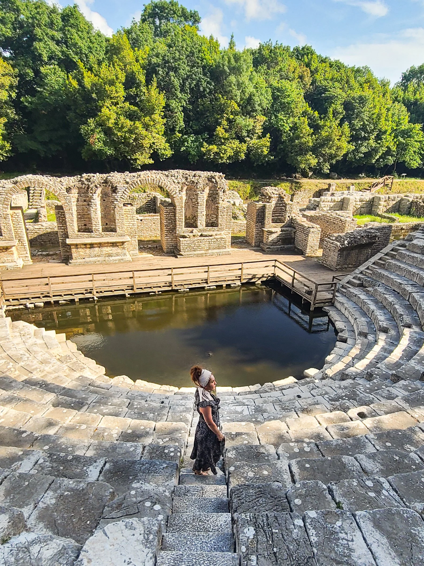 An ancient Roman theater with stone steps, a small reflecting pool, and ruins of arches, surrounded by green trees and a partly cloudy sky. A woman in a black dress and headband is walking on the steps.